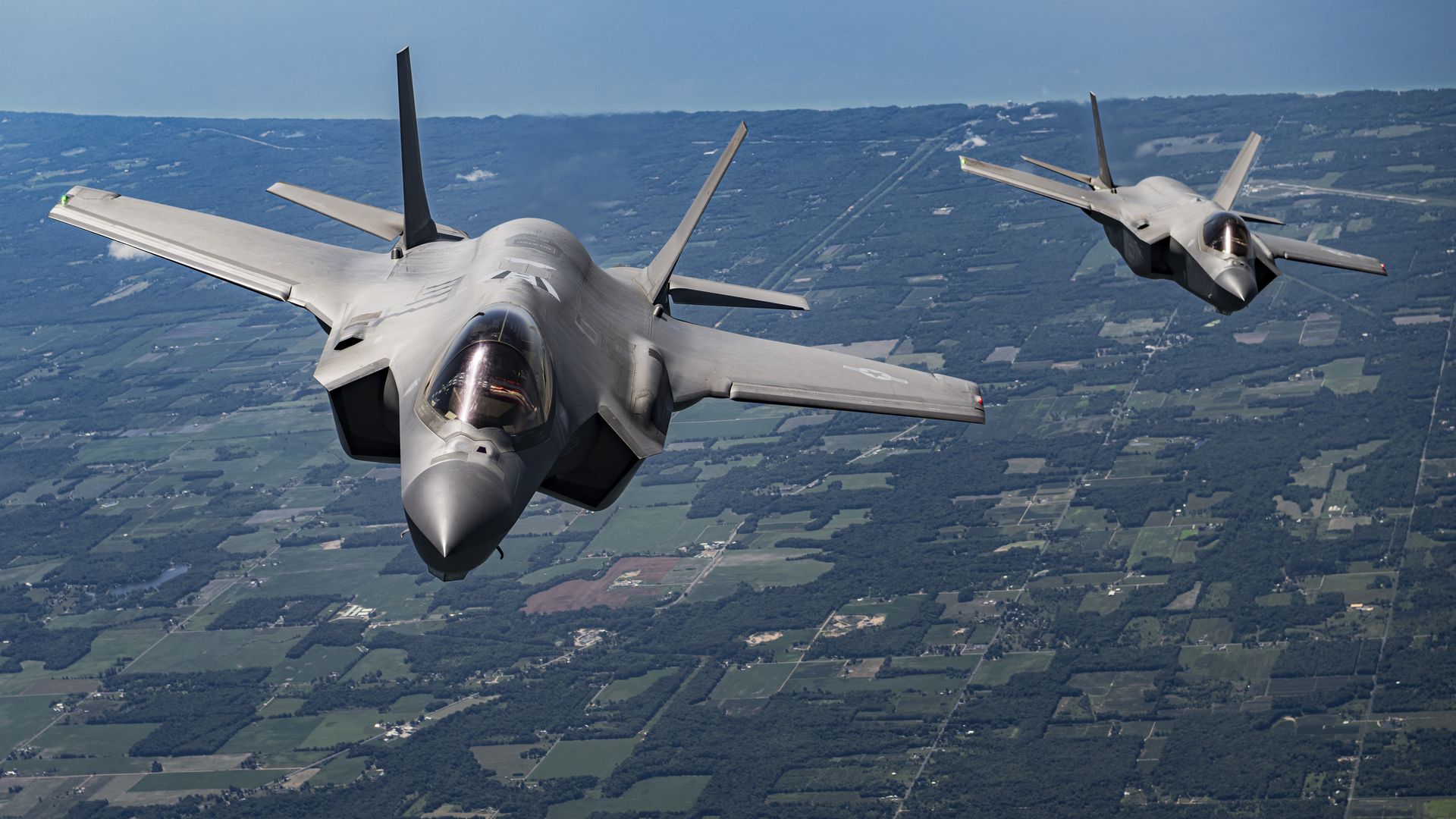 Two gray fighter jets flying over green farmland and forested areas under a clear blue sky.