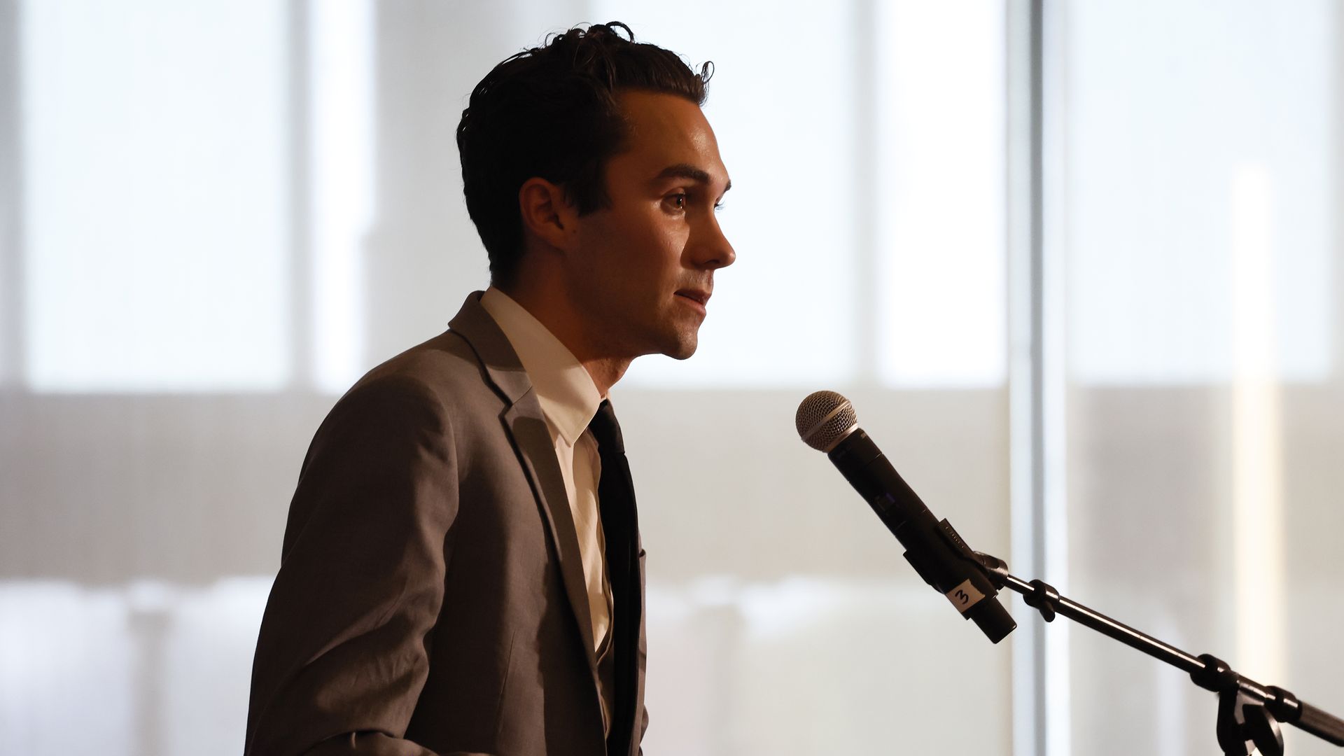 David Hogg, wearing a gray suit and speaking into a microphone in front of a glass wall.