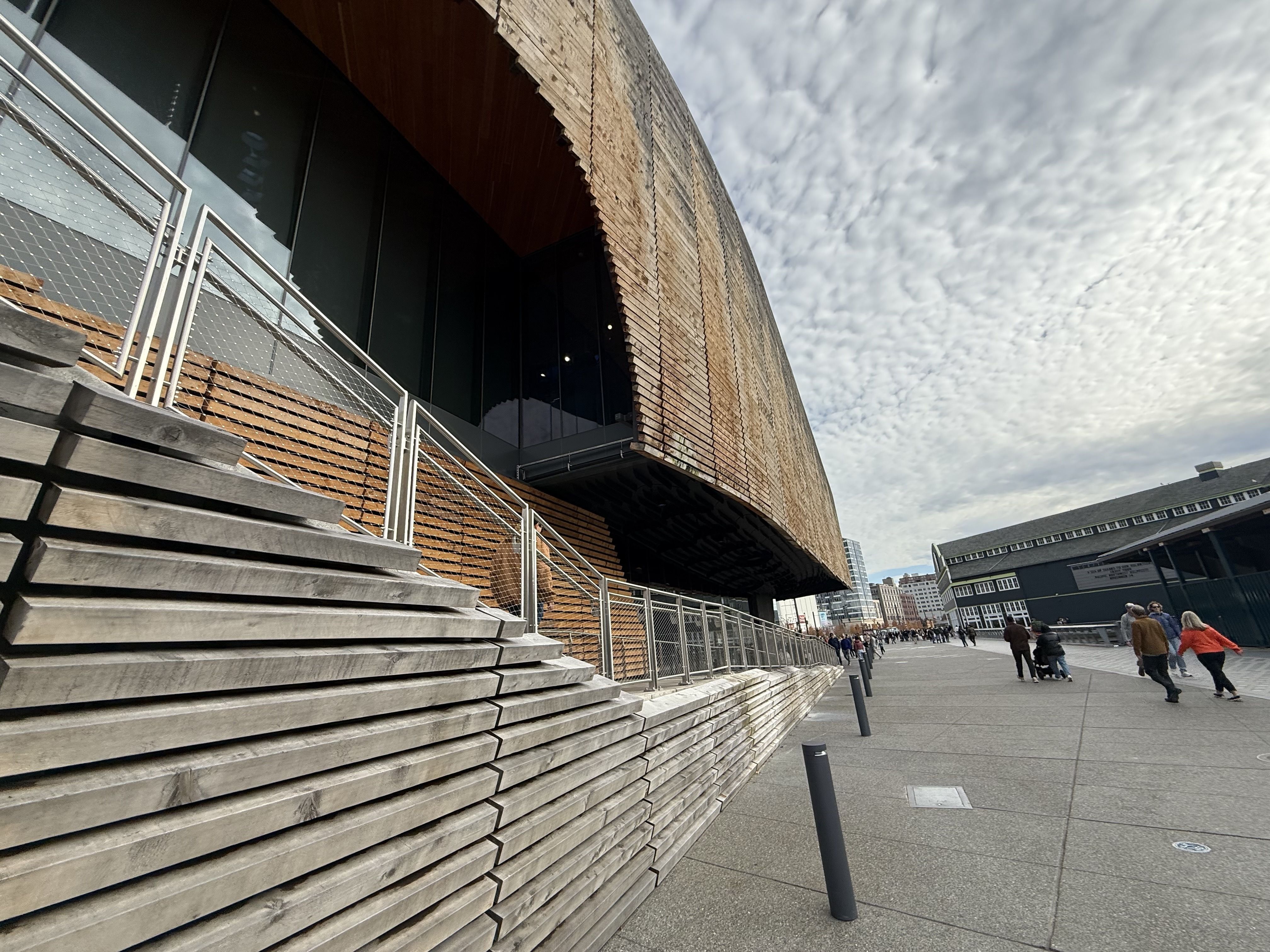 Modern building with angled wooden slats and glass entrance on the left; pedestrians walk on a wide sidewalk under a cloudy sky.