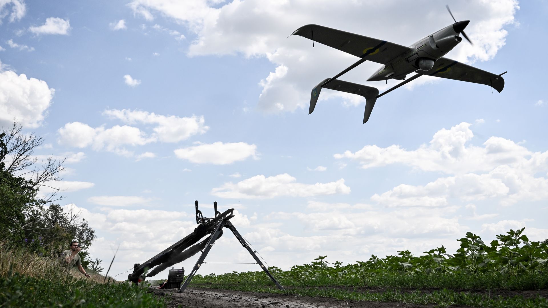 A Ukrainian drone launches from a rail in a green field. A military member can be seen squatting in the left corner. The sky is blue.