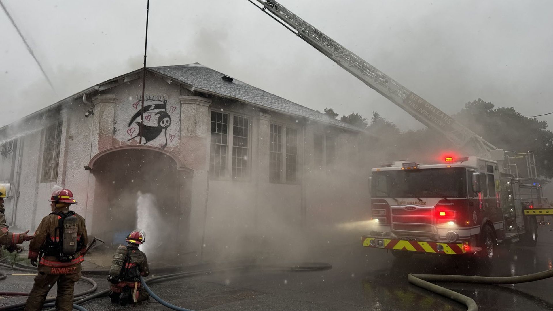 Firefighters work to put out a fire in a white building. One kneels down and aims a fire hose into the front door.