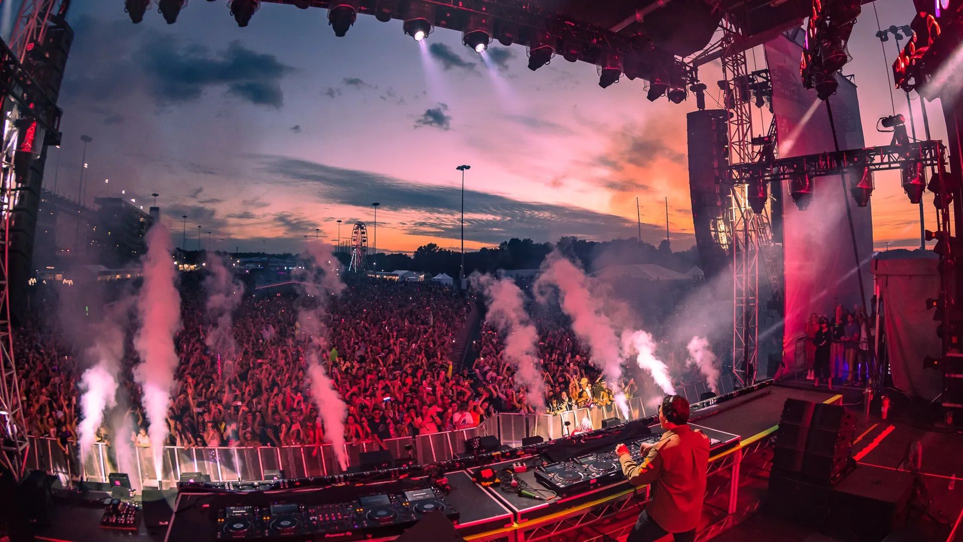 DJ performing on stage at sunset with colorful lights and smoke effects, facing a large lively crowd enjoying the outdoor music festival, with a Ferris wheel visible in the background.