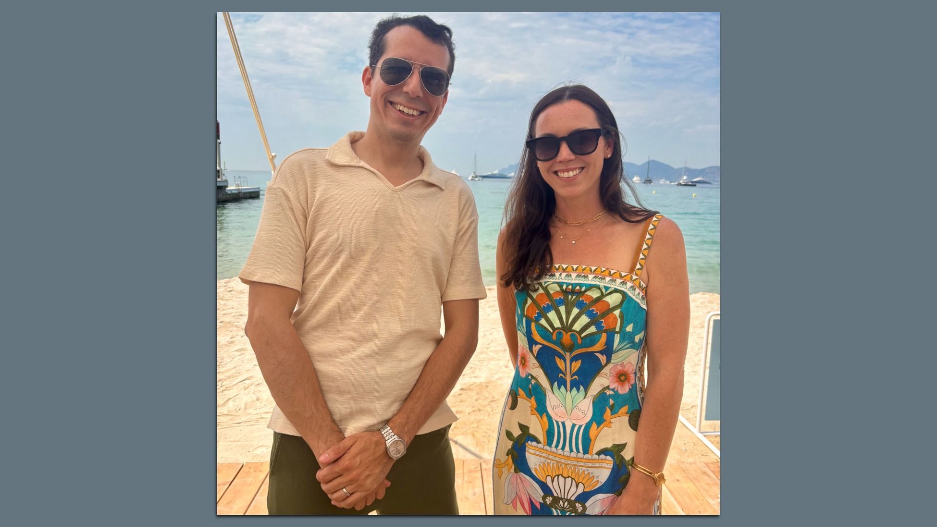 Simon Hernandez-Arthur and Eleanor Hawkins wearing sunglasses smile on a sunny beach; Hernandez-Arthur in a beige polo and Hawkins in a colorful patterned dress, with turquoise water and sailboats in the background.