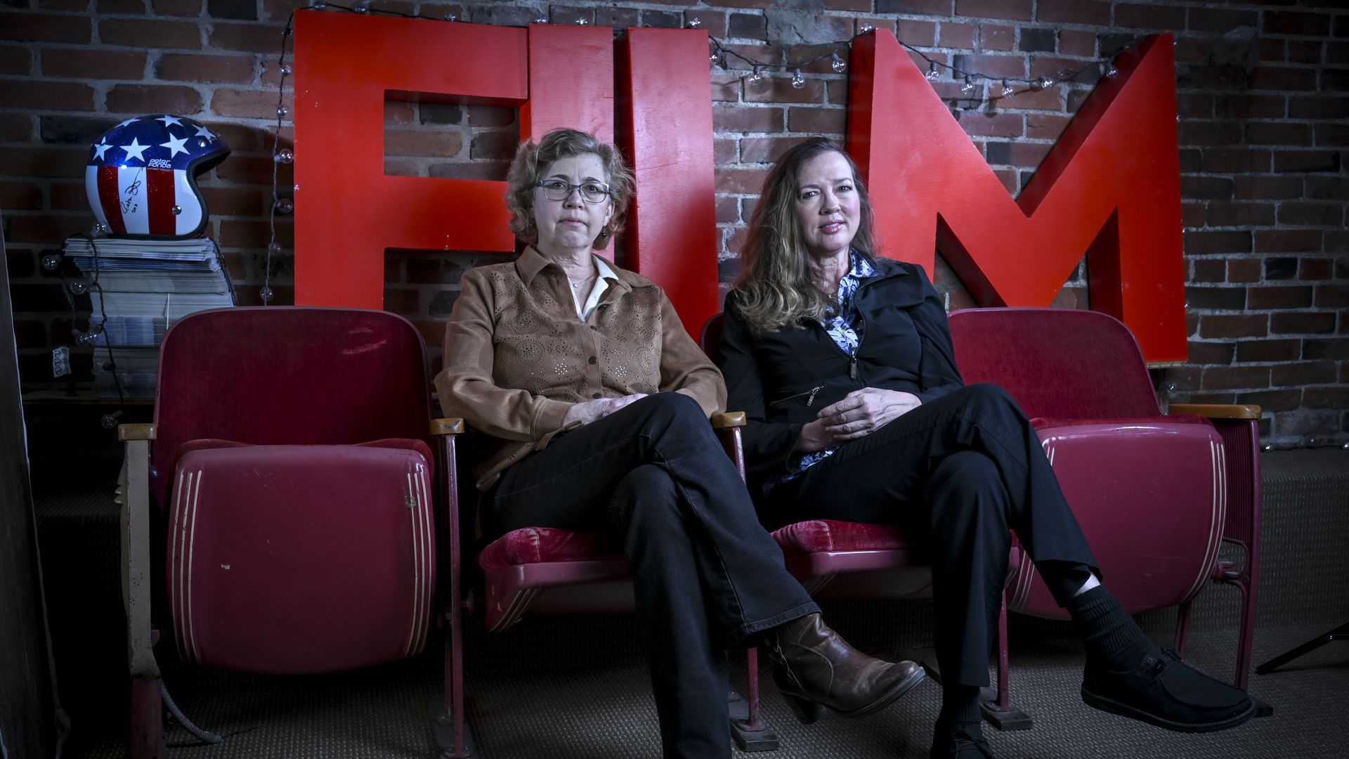 Boulder International Film Festival co-founders Kathy and Robin Beeck sit on red theater seats in front of a brick wall and large red letters behind them. A patriotic blue helmet with white stars sits on a stack of papers to the left; string lights hang above.