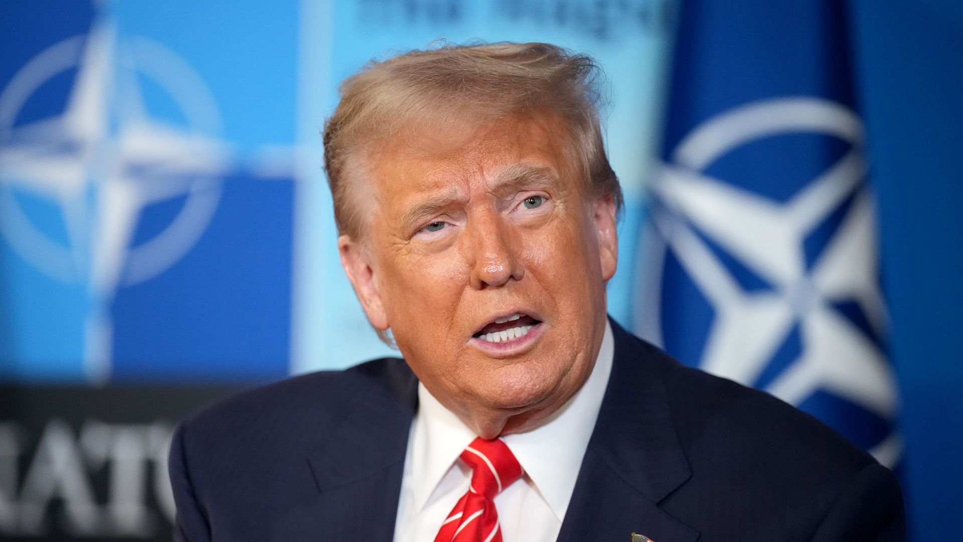 President Trump in a dark suit and red striped tie speaks in front of a backdrop featuring the NATO logo. He wears an American flag pin on his lapel.