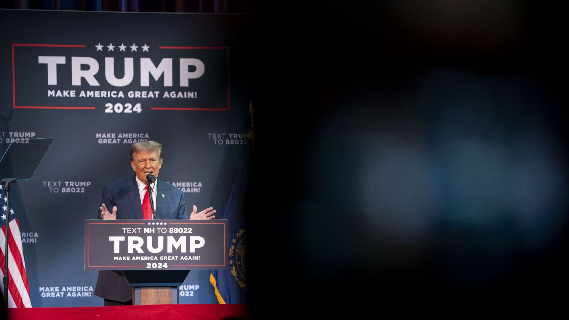 Donald Trump speaks behind a podium that says "Trump," wearing a blue suit, white shirt and red tie.