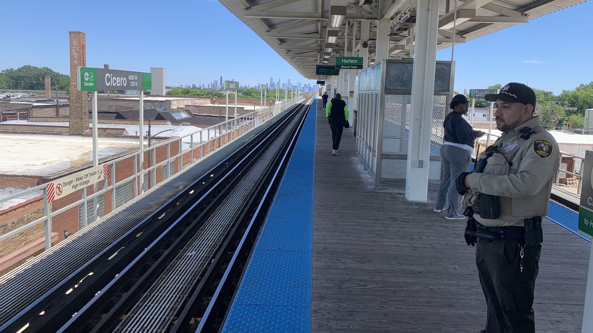 An elevated subway platform with blue textured edge and steel beams. Tracks run center; signs read "Cicero" and "Harlem." A police officer stands at the right, skyline in distance under a blue sky.
