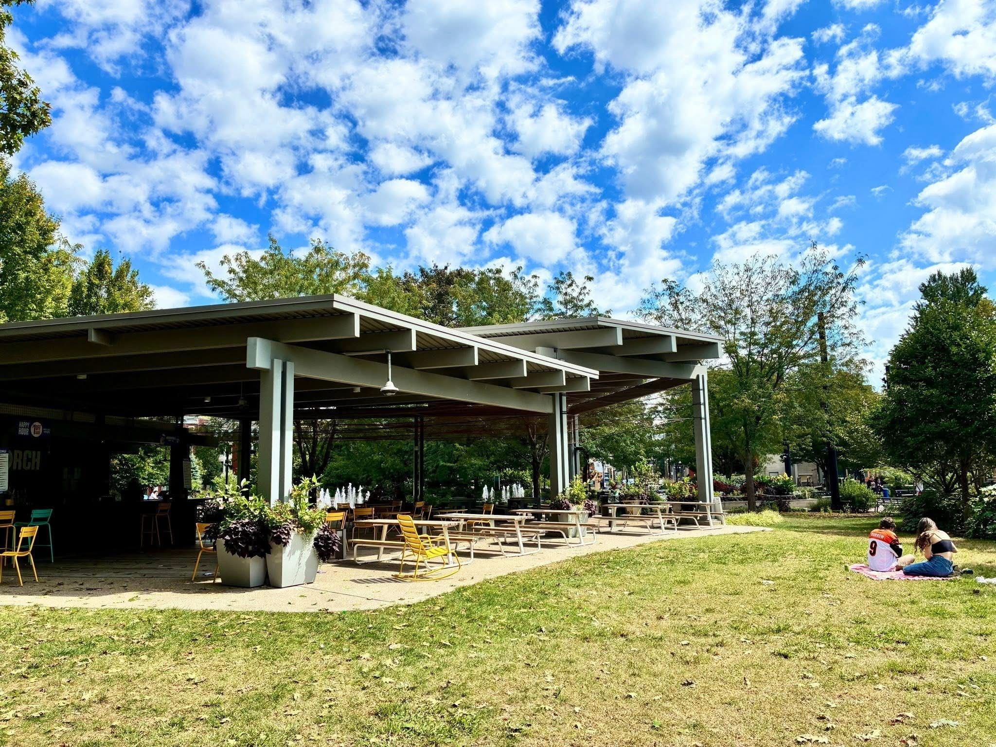 Blue sky with scattered white clouds above an open-air pavilion. Gray structure with yellow chairs and potted plants; people sit on a blanket on the grass to the right.
