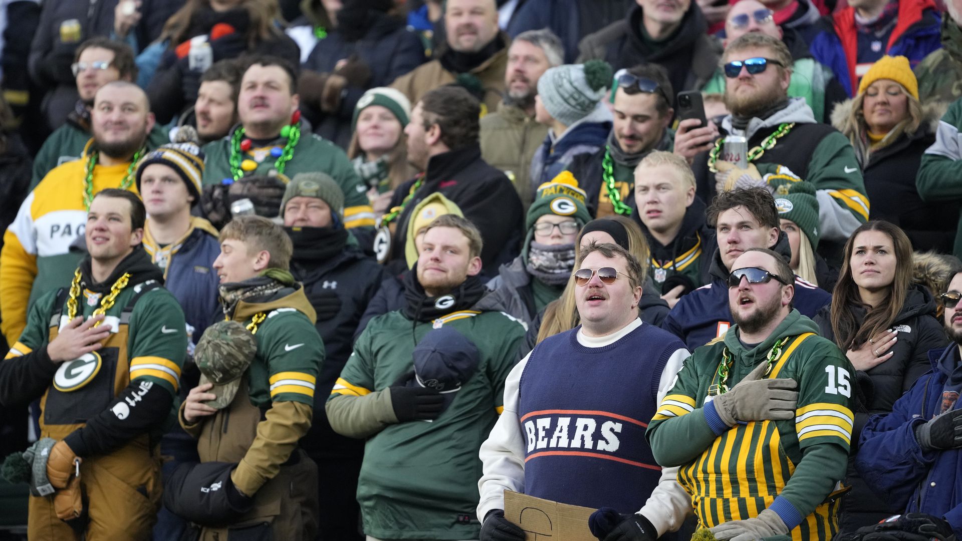 Crowd of fans at an outdoor football game, many wearing green and yellow Green Bay Packers gear, some with hands over hearts, one man in a blue Bears sweater, cold weather attire.