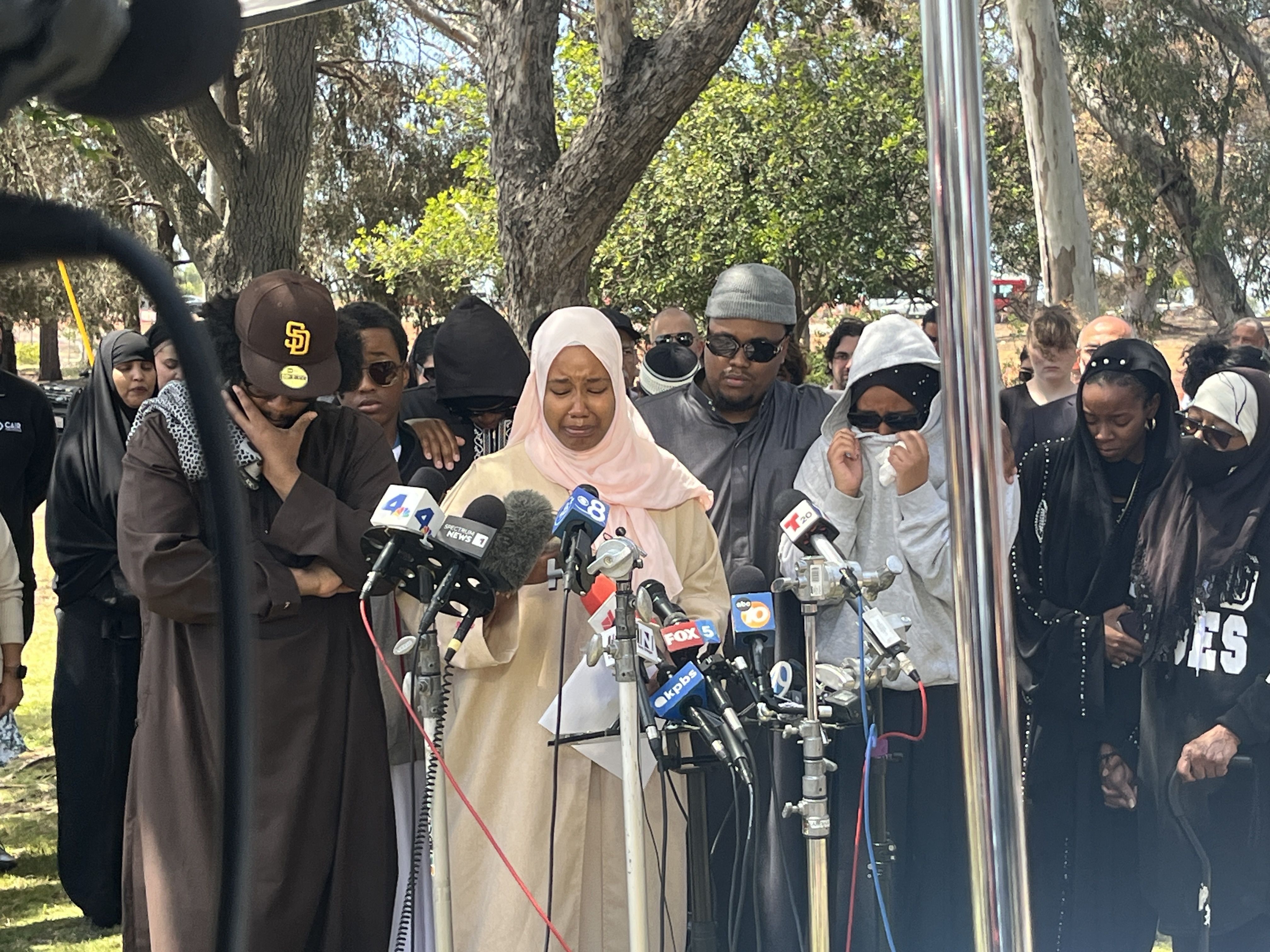 Outdoor press conference; a woman in a pale pink hijab speaks into multiple microphones, surrounded by reporters and attendees in hijabs, with trees in the background.