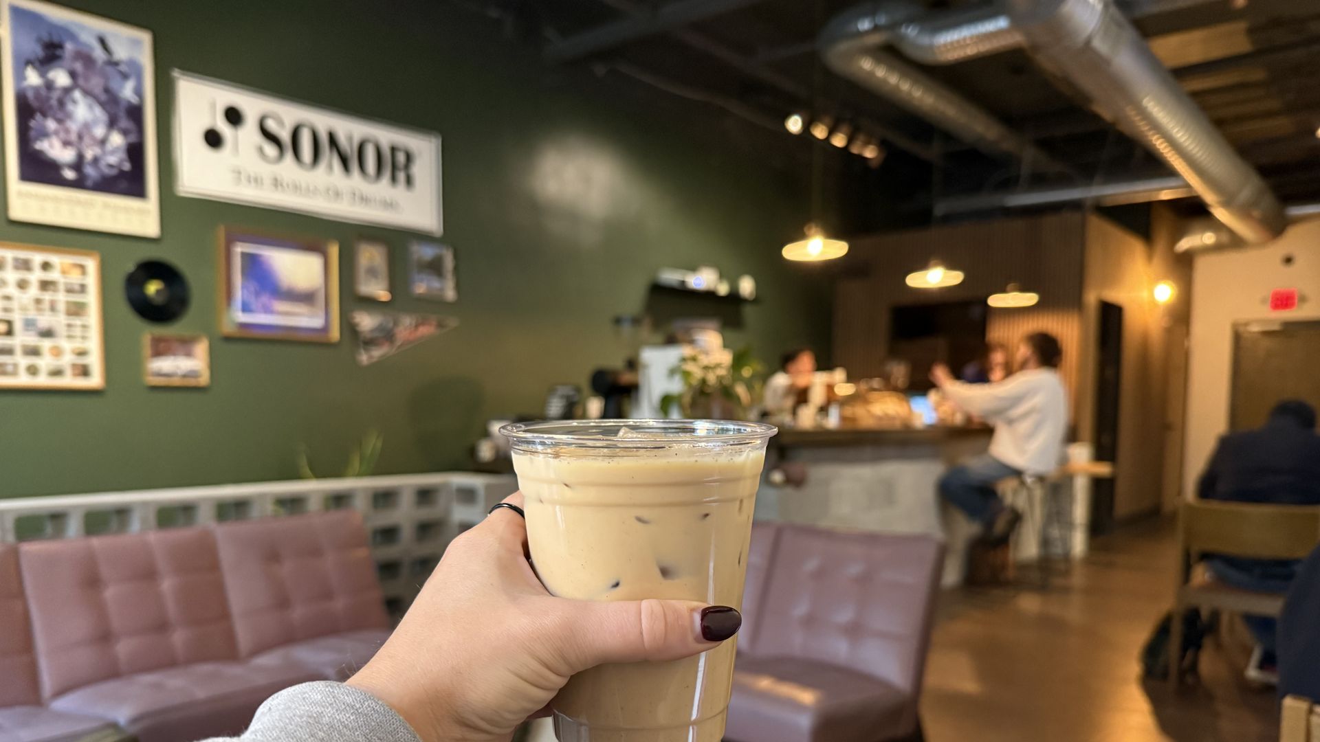 Hand holding a plastic cup of iced coffee inside a cozy cafe with mauve chairs, green walls decorated with framed pictures, and people sitting at a wooden counter under warm lights.