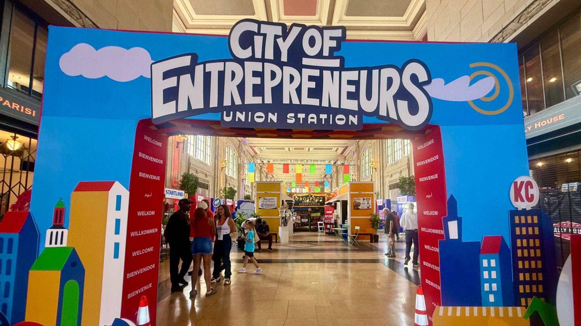 Colorful expo entrance with a blue arch reading "City of Entrepreneurs Union Station". Red welcome pillars flank the aisle; people walk toward vendor booths beneath banners inside a mall.