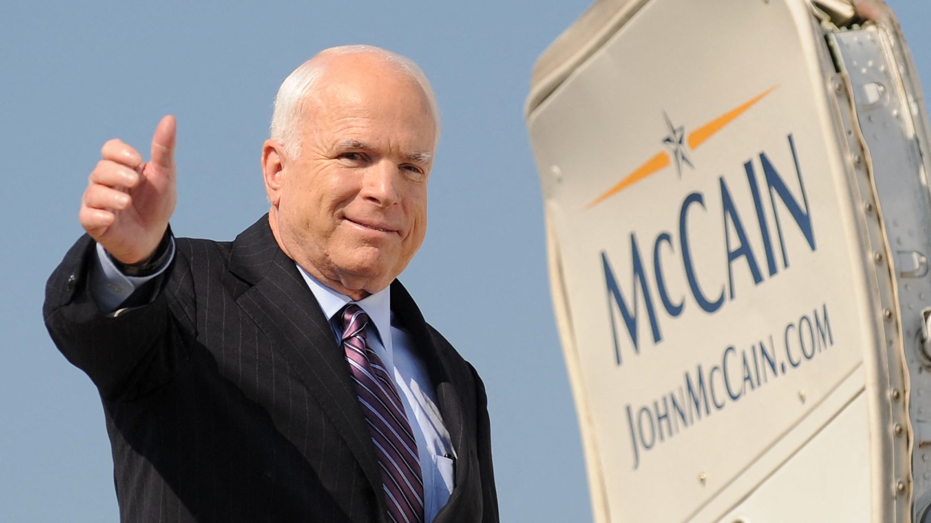 A smiling man gives a thumbs up next to an open airplane door with the word McCain written on it.