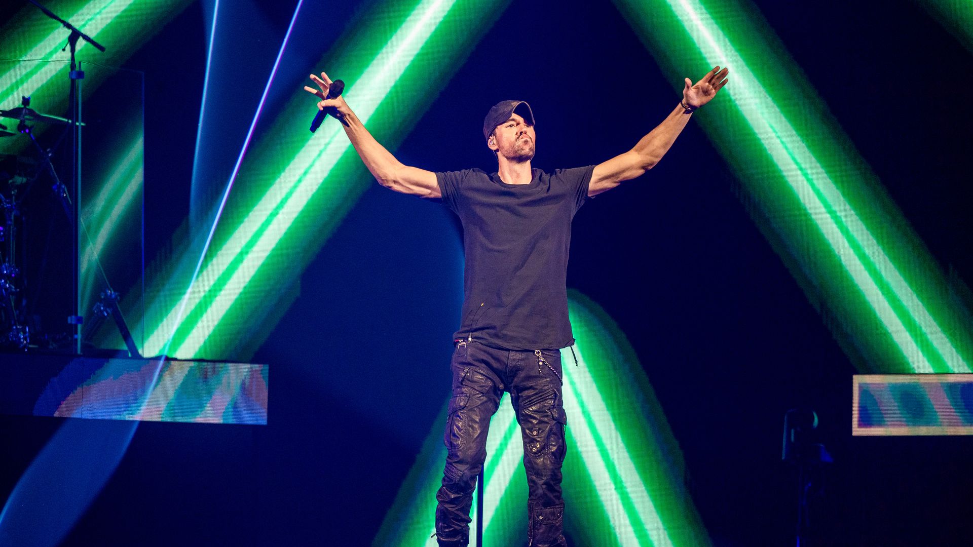 A super good-looking man in black shirt and pants raises his arms while laser lights flash behind him