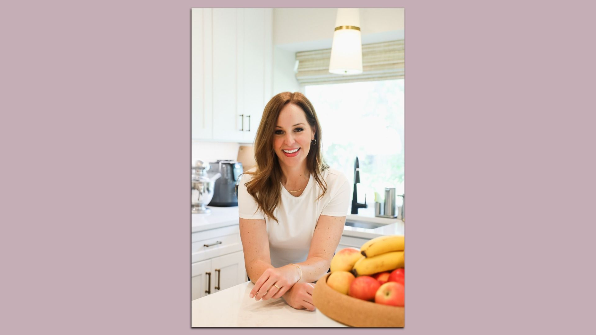 Smiling woman in a white shirt leans on a kitchen island, with a bowl of apples and bananas in the foreground. White cabinets, a coffee machine, and a pendant light visible in a bright kitchen.