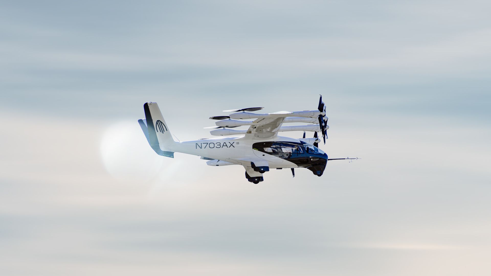 White and blue tilt-rotor aircraft with rotors on its wings, flying across a pale blue sky; registration N703AX on the fuselage.