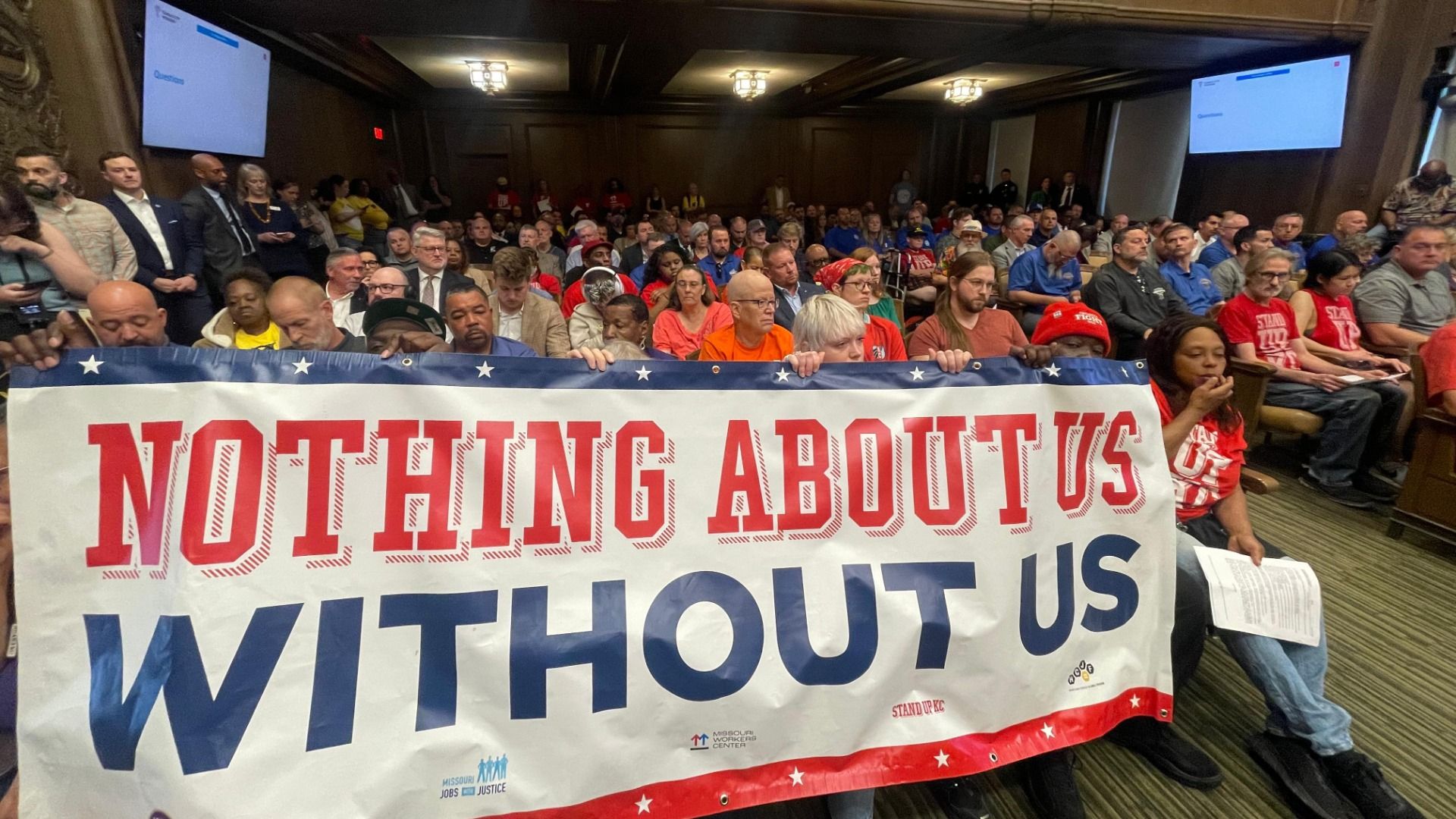 Crowded conference room with attendees sitting and standing behind a large banner that reads "NOTHING ABOUT US WITHOUT US" in red and blue, featuring white stars; many wear red shirts.