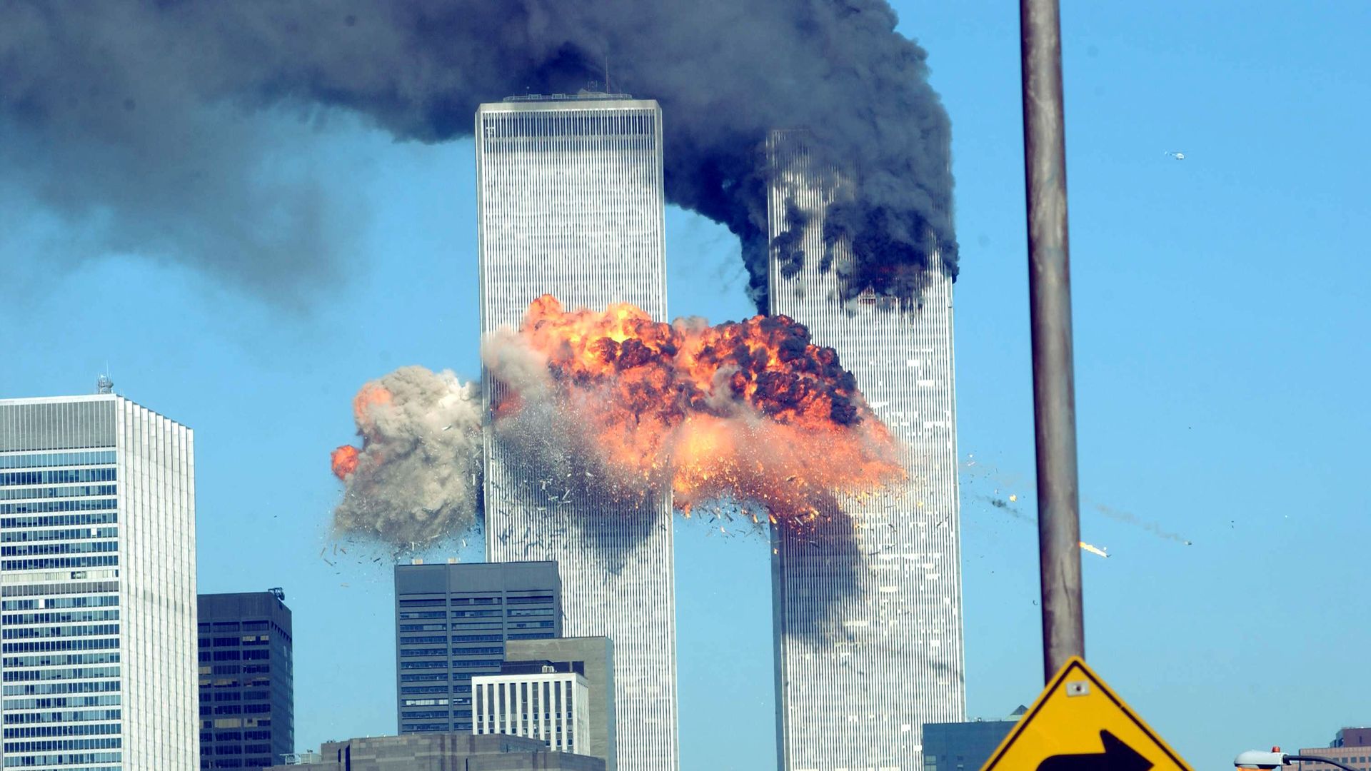 Explosions and thick black smoke billowing from the World Trade Center Twin Towers in New York City against a clear blue sky with city buildings and a right-turn traffic sign in the foreground.