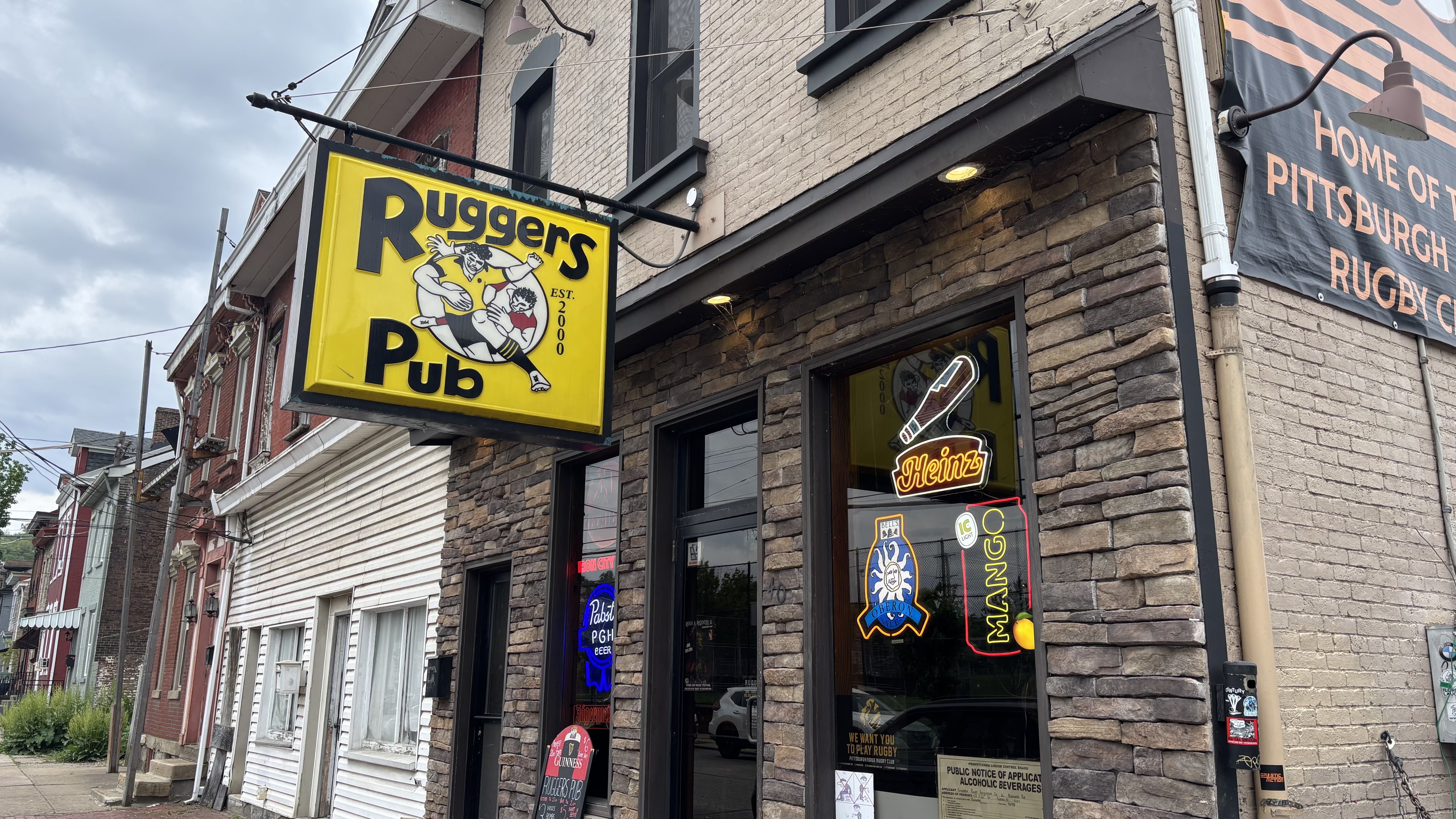 Bright yellow pub sign for Rugger's Pub with a cartoon rugby player on a brick storefront; neon Heinz sign in the window; banner reading Home of Pittsburgh Rugby with orange stripes; cloudy sky.