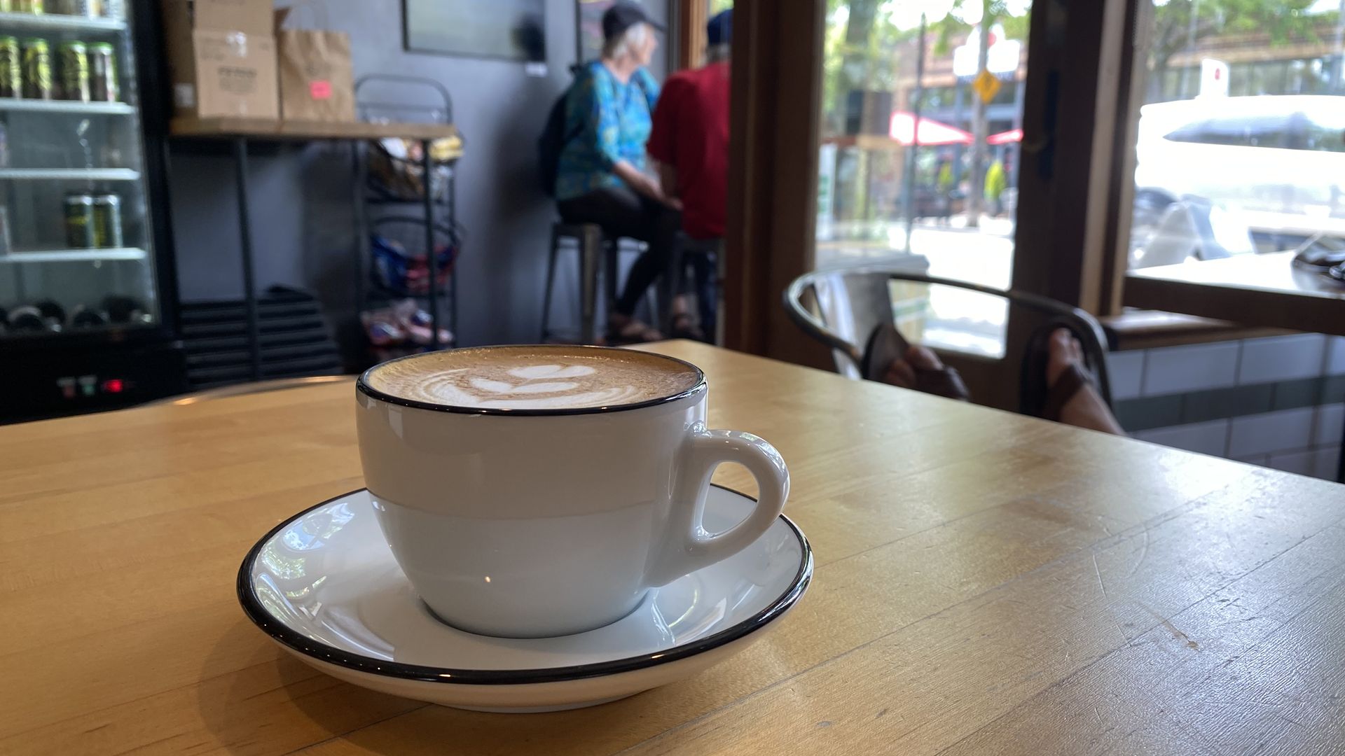 A cappuccino with decorative foam art on in a porcelain cup and saucer on a wooden table, with people talking and windows in background.