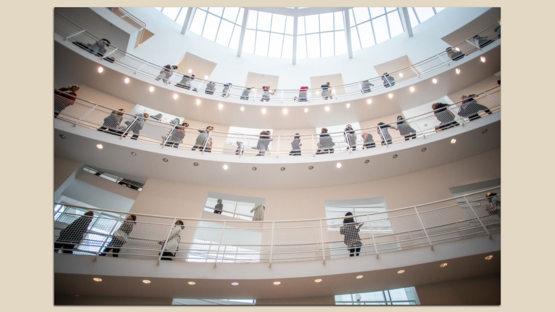 A spacious, white, multi-level circular atrium with curved balconies. Numerous people stand along the rails on every level, looking into the open central space beneath a large skylight.