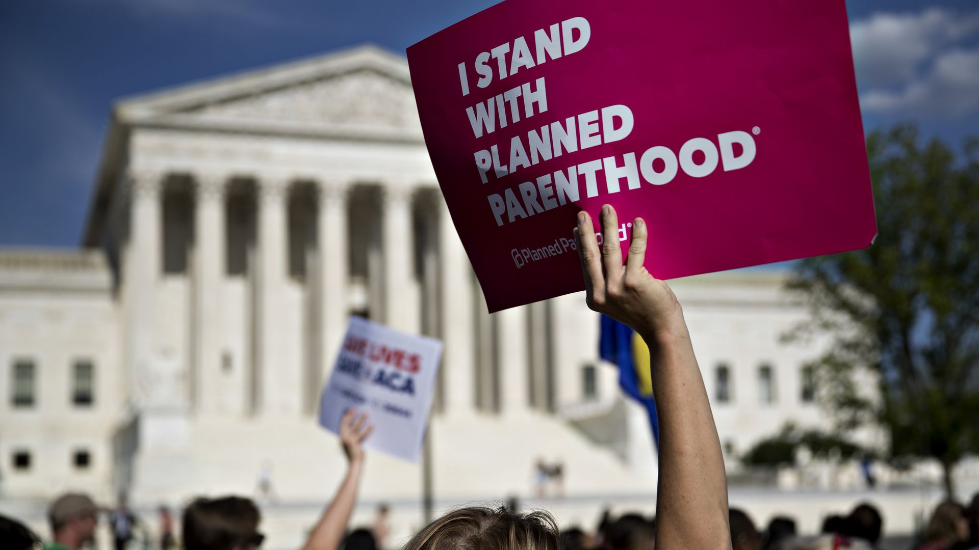 Picture of a hand holding a pink sign that says "I stand with Planned Parenthood" in an abortion rights protest happening in front of the U.S. Supreme Court