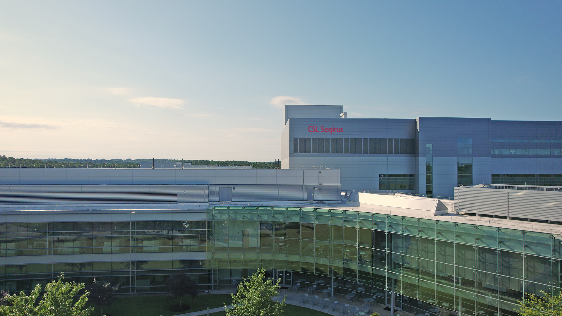 Modern corporate building with gray panels and a large glass facade, red CSL Seqirus sign on the wall, clear blue sky, and distant tree line on the horizon.