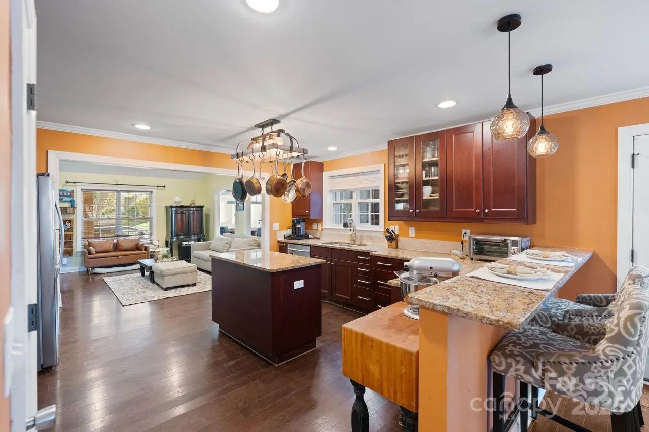 Bright kitchen with orange walls, dark wood cabinets, granite countertops, hanging pots, a small island, and two patterned bar stools. Adjacent living room with leather and fabric sofas.