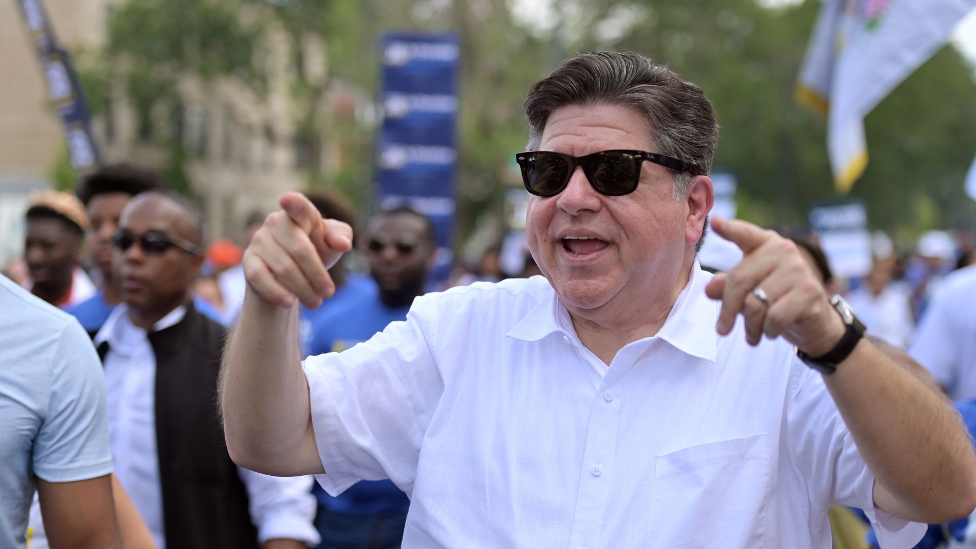 JB Pritzker, in sunglasses and a white short-sleeve shirt, smiles and gestures.