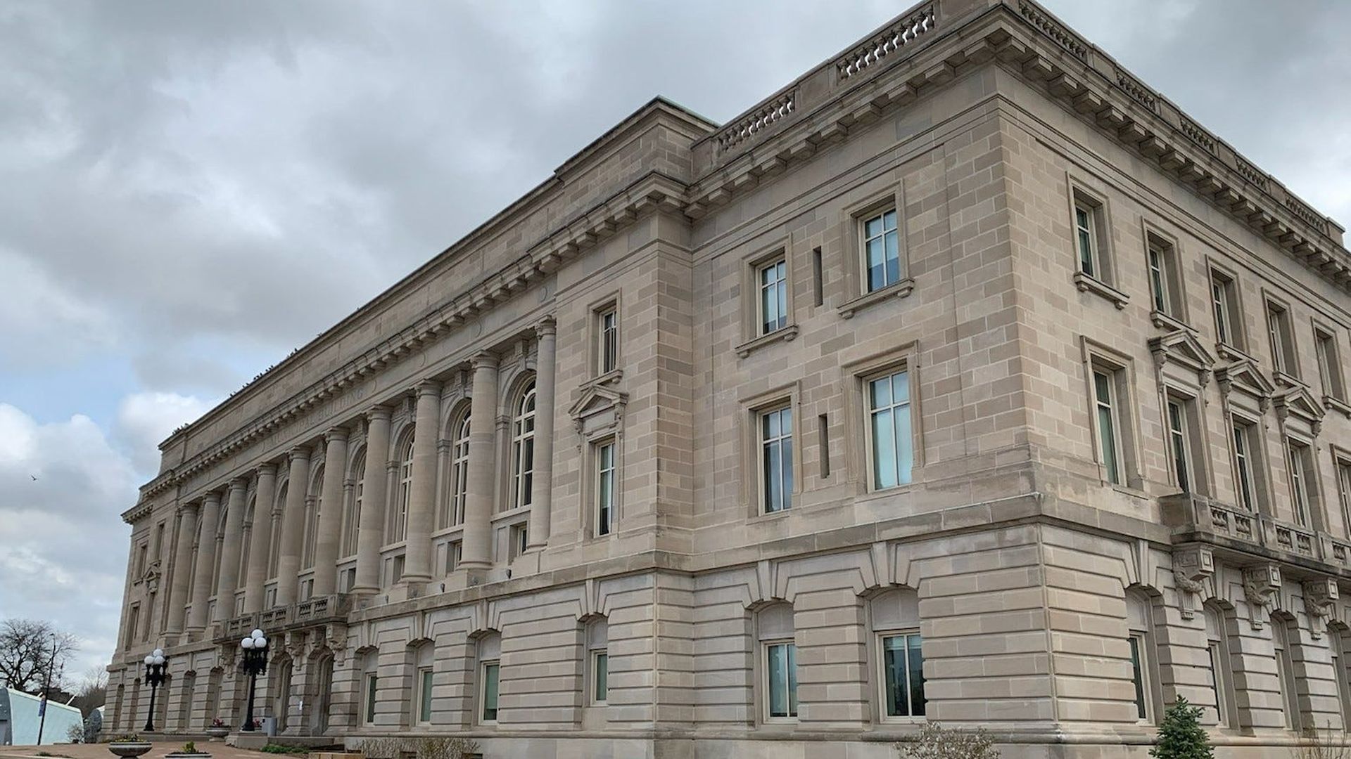 Beige stone neoclassical building with tall arched windows along the left, rectangular upper windows, and ornate cornice. A paved plaza with planters and black lampposts sits in front under a cloudy sky.
