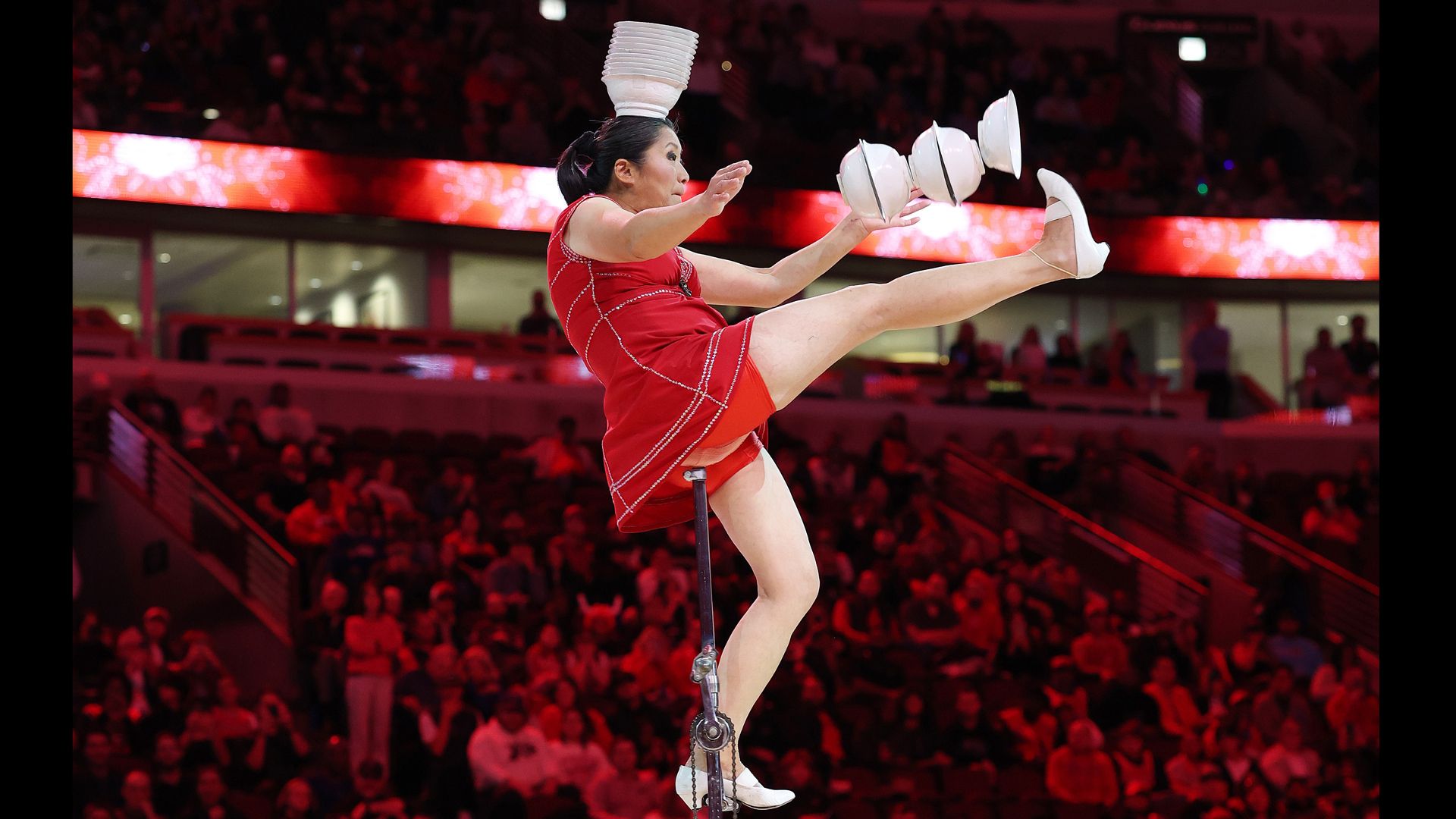 A woman in a red dress on a unicycle with bowls on her head and hands