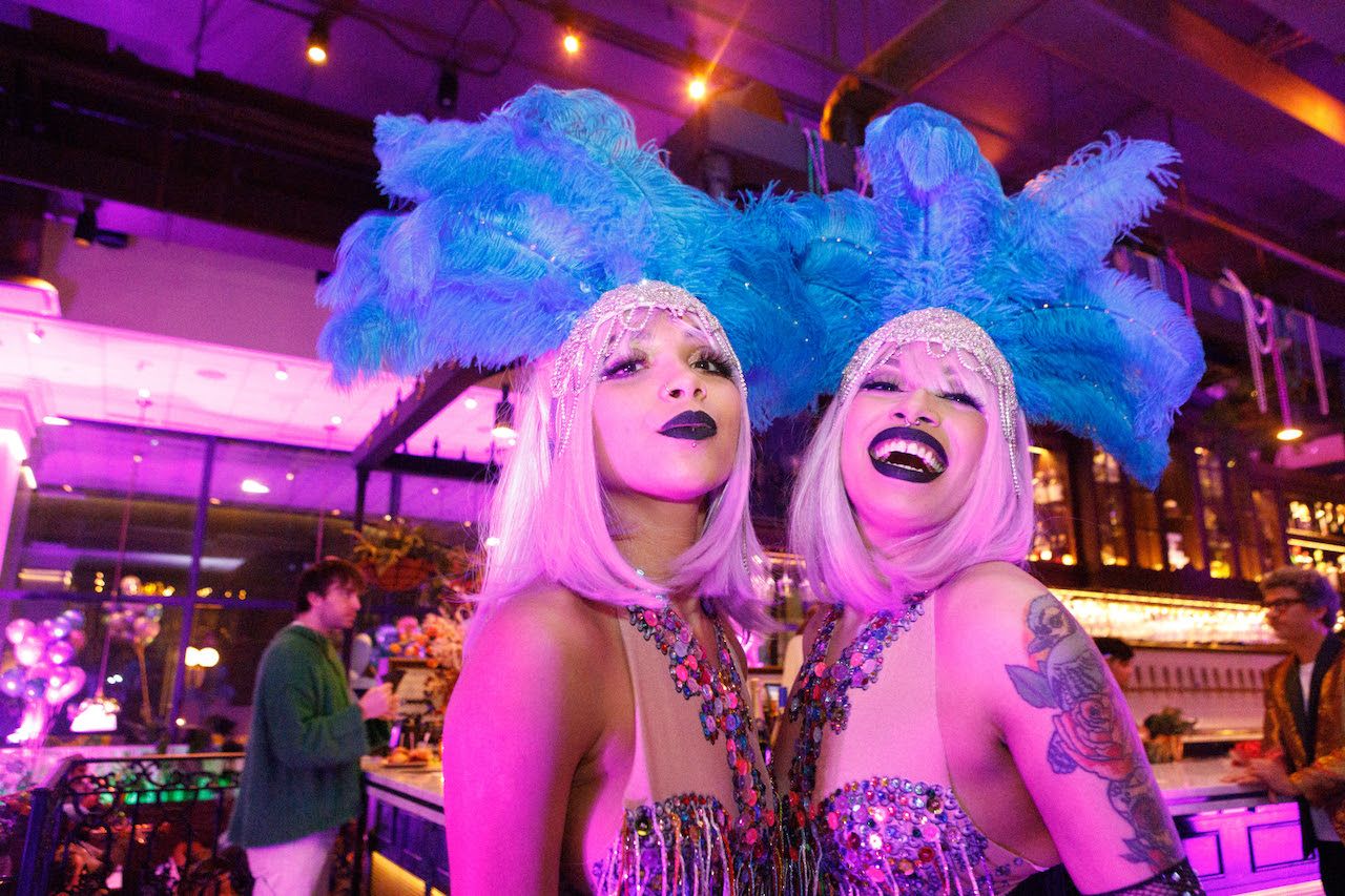 Two women with platinum blonde wigs and large blue feather headpieces wear sequined costumes and dark lipstick, smiling inside a purple-lit bar or club with people in the background.