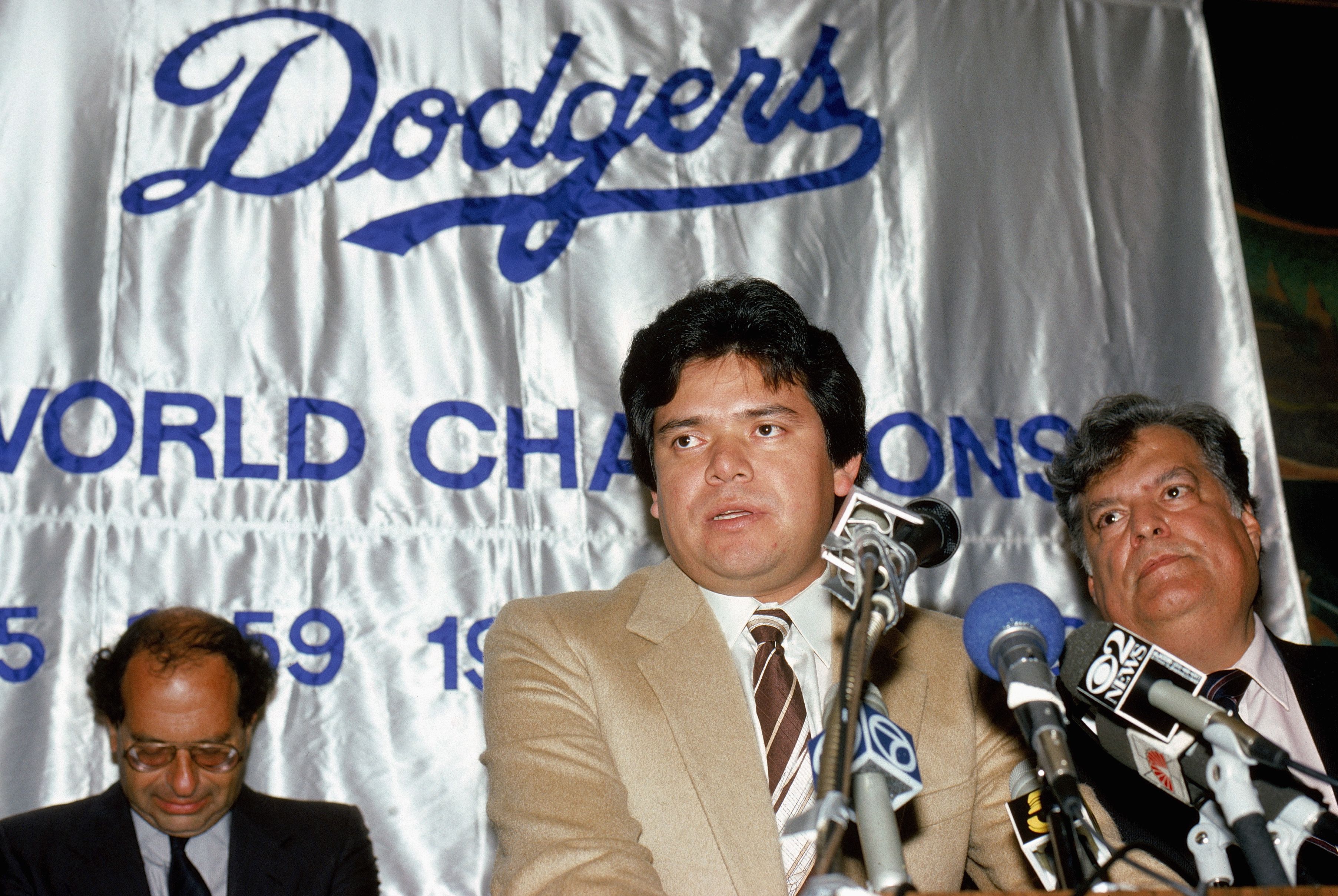 Fernando Valenzuela, in a tan suit, speaks in front of several microphones during a press conference.