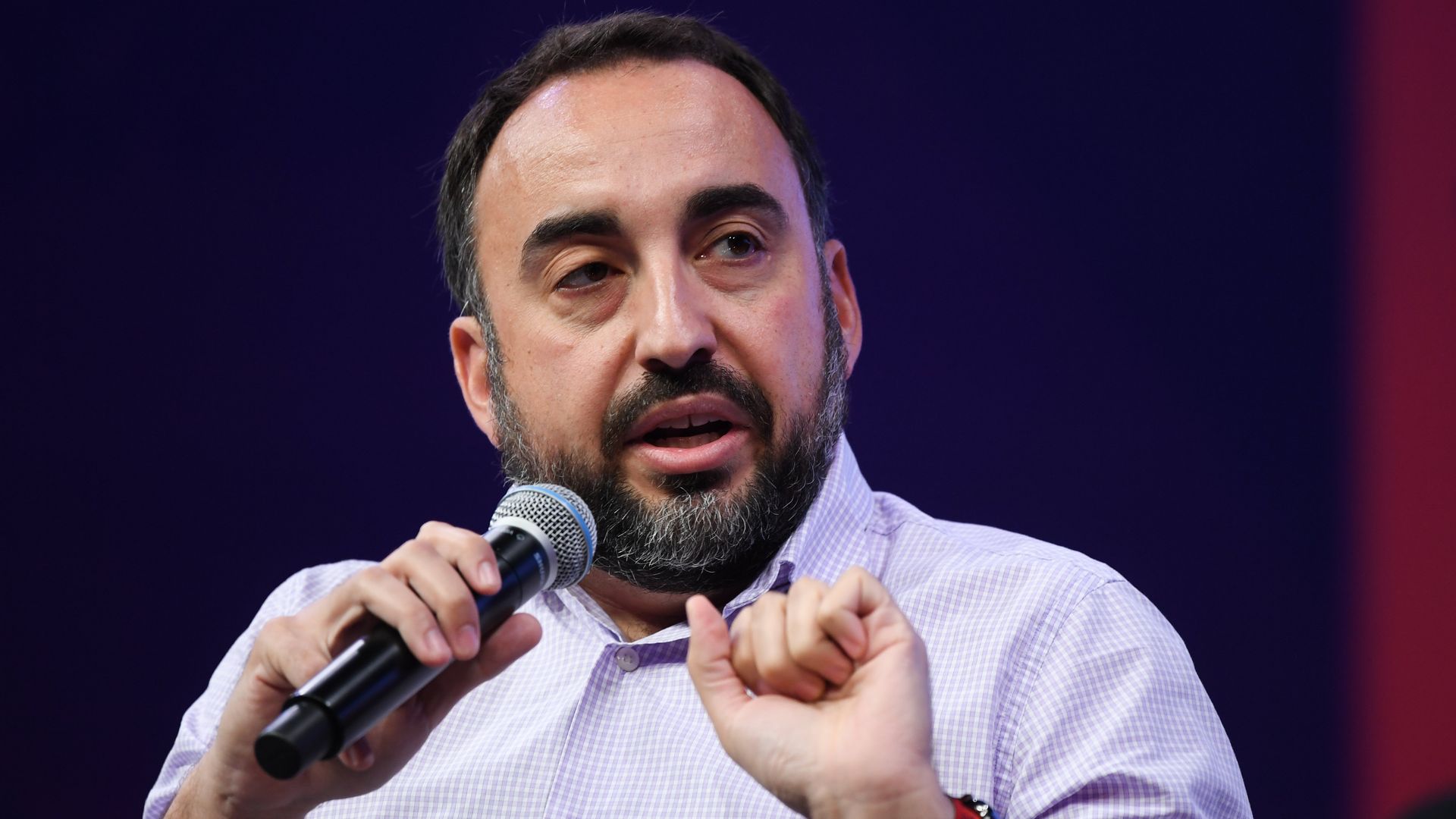 A man with black hair and a beard talking into a microphone and wearing a purple checkered shirt.