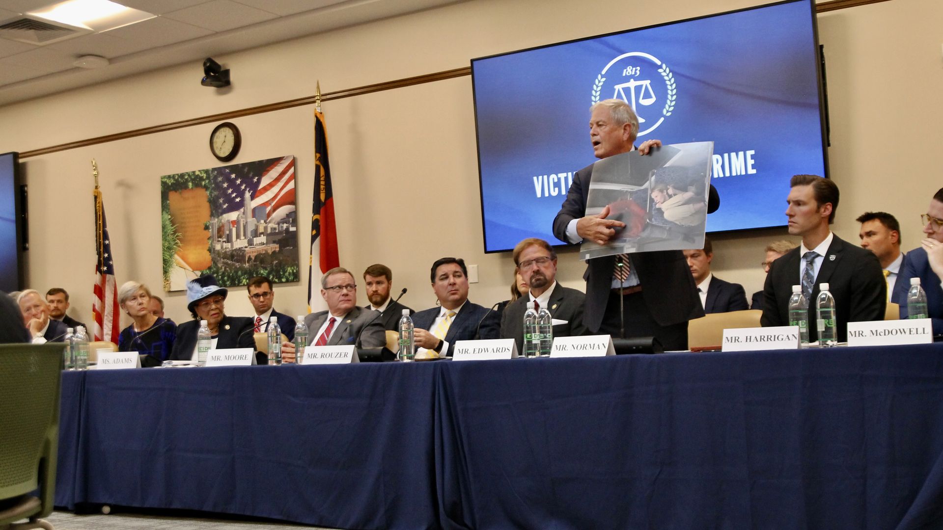 A formal meeting with a panel of men and women seated behind a table with nameplates in front, a man standing presenting an image, American and North Carolina flags in the background.