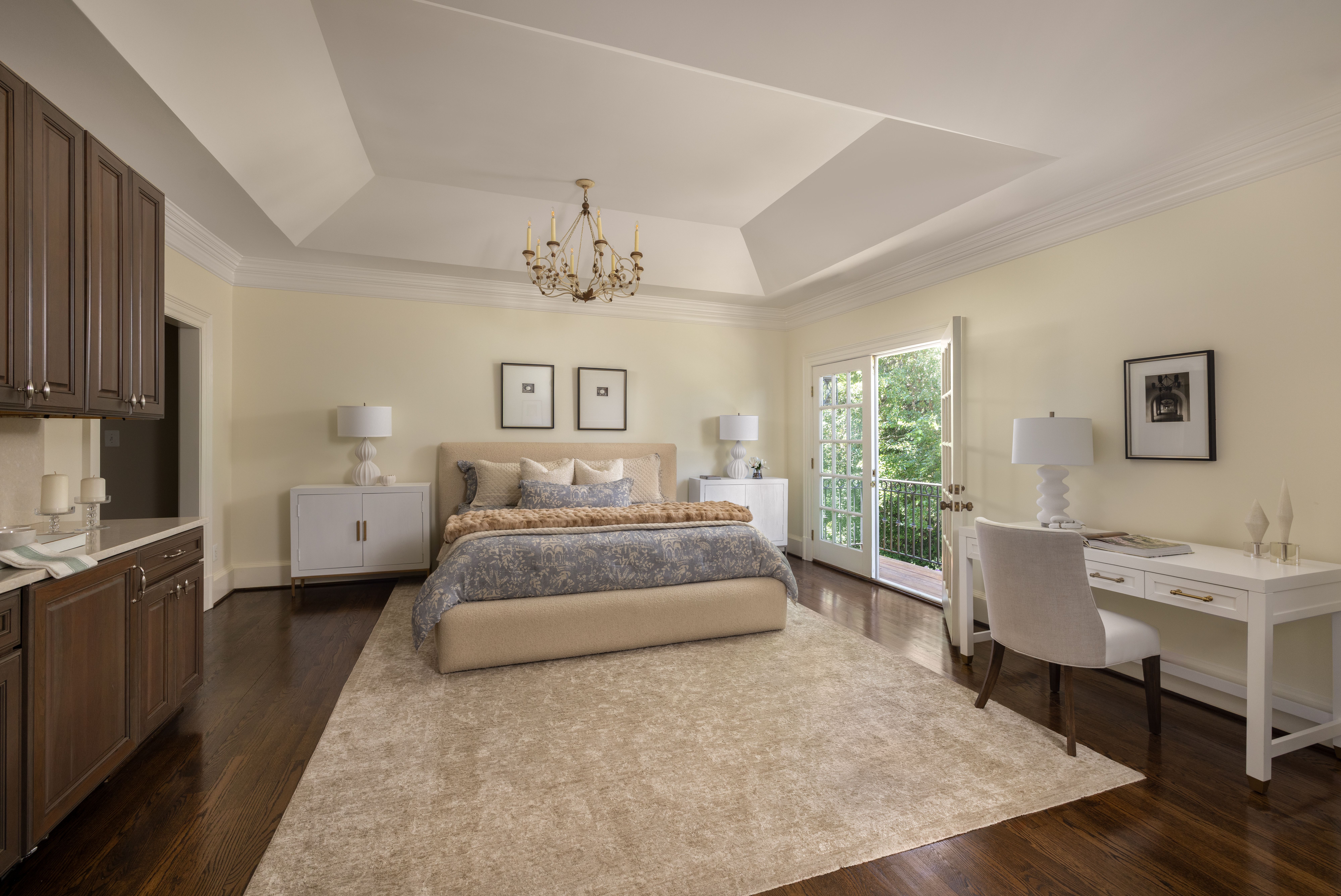 A spacious beige bedroom with a central chandelier, beige bed with blue bedding, dark wood floors, white nightstands with lamps, a desk, framed art, and French doors to a green outdoor view.