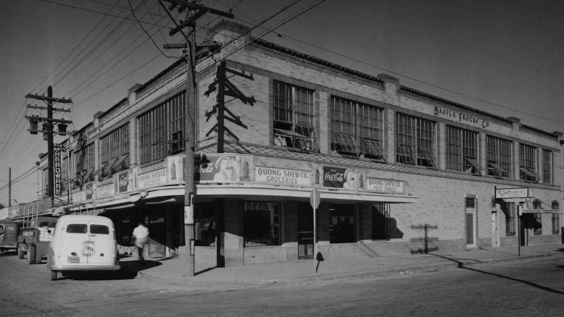 A black-and-white photo shows a two-story, brick building on a street corner with vintage cars parked outside.