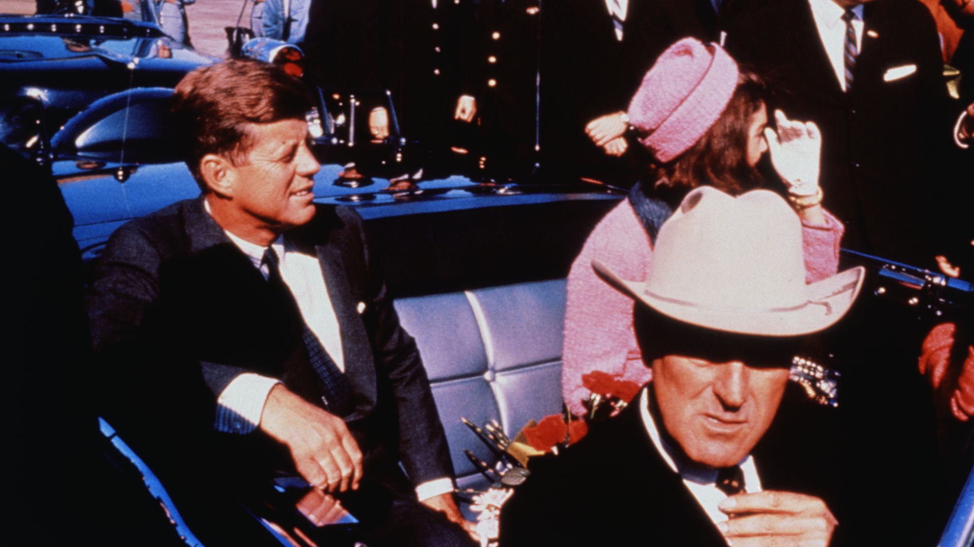 Texas Governor John Connally adjusts his tie (foreground) as US President John F Kennedy (left) & First Lady Jacqueline Kennedy (in pink) settled in rear seats, prepared for motorcade into city. 