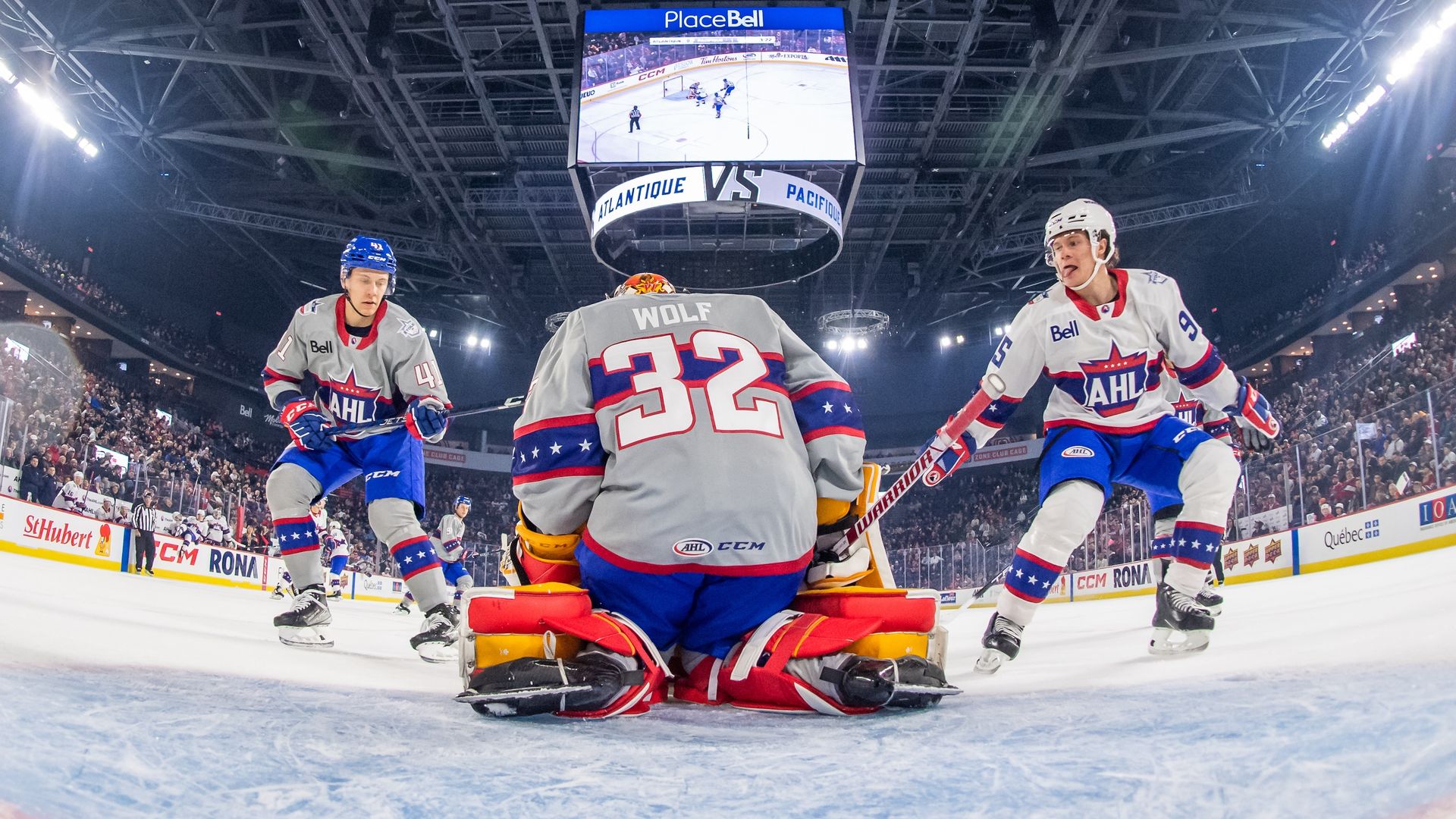 Ice hockey game at Place Bell arena with players wearing gray and white jerseys with red and blue stars. Goalkeeper number 32 named Wolf kneels in front of goal as two players flank him.