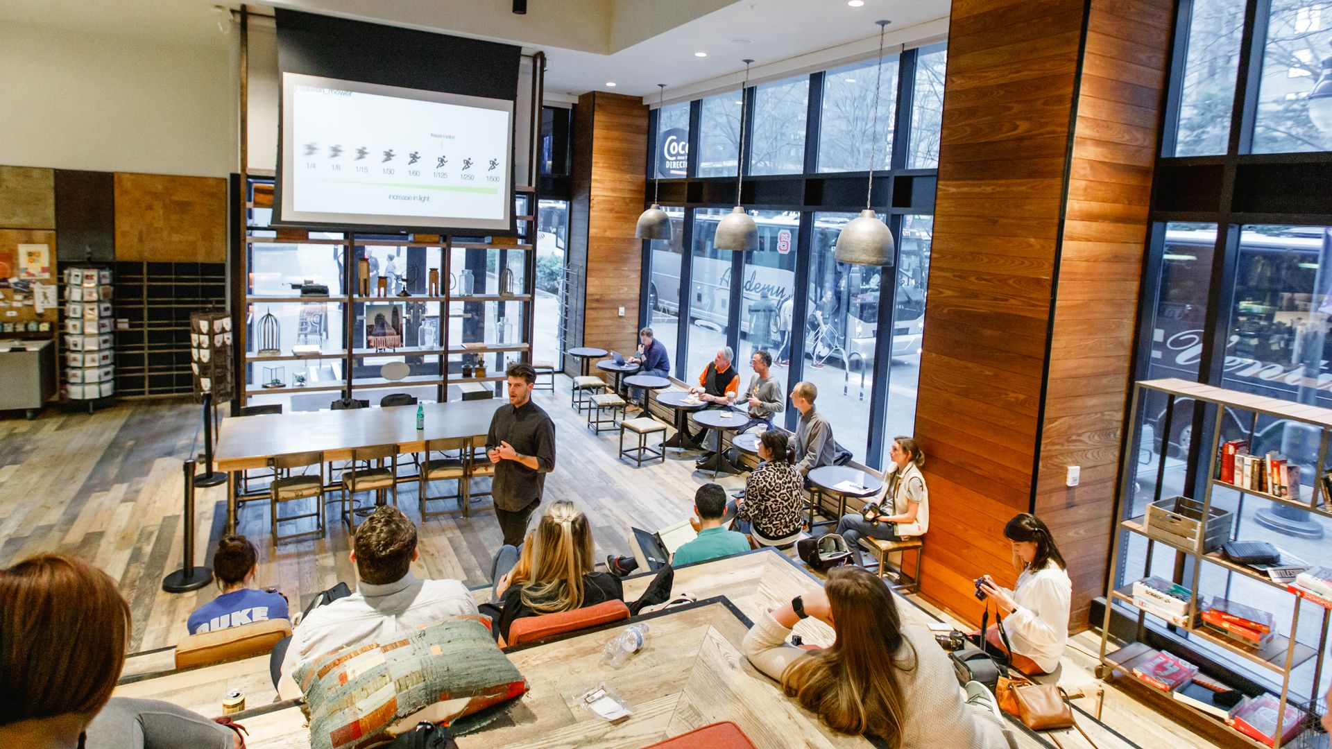 Modern classroom with wooden floors and large windows, a man presents photography shutter speed slides to students sitting on tiered wooden benches and small round tables.
