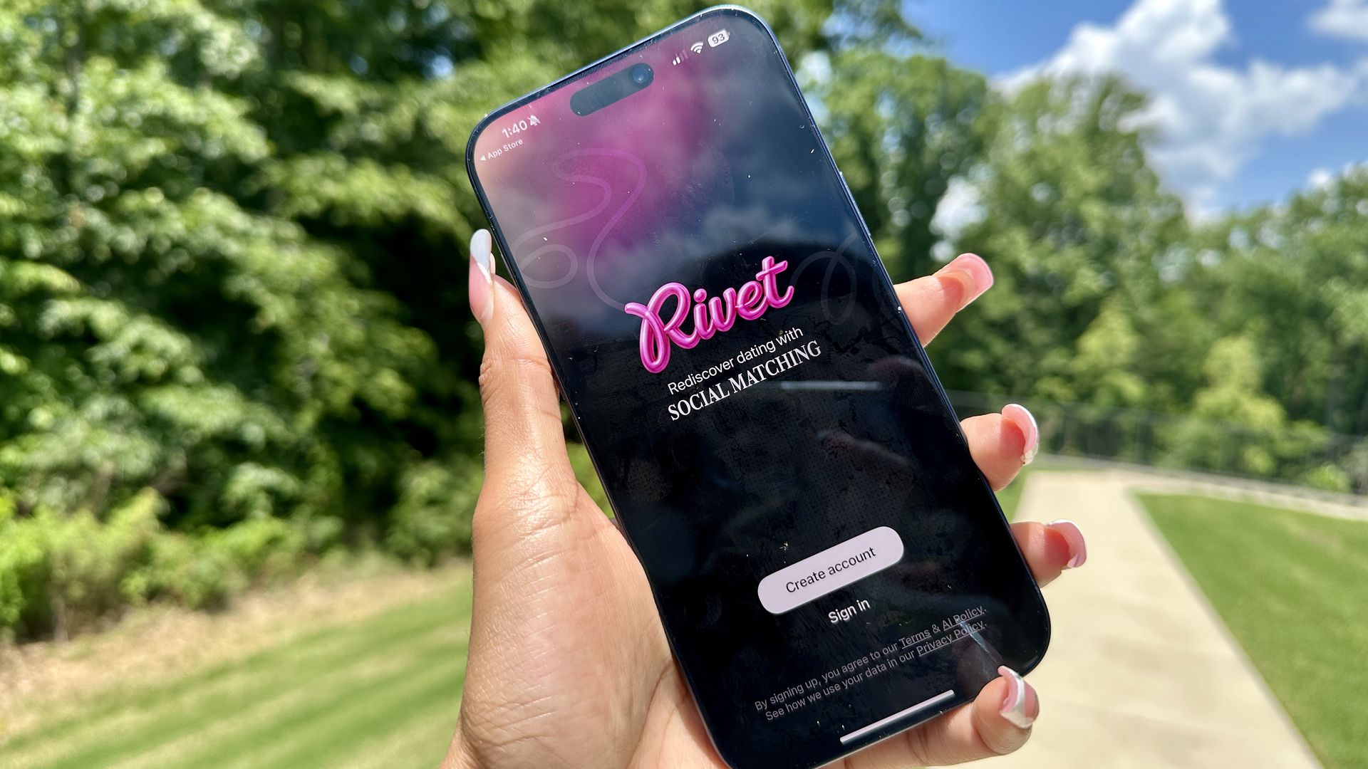 Rivet dating app. Person holds a smartphone outdoors; the screen shows a dating app with the pink logo "Rivert" and the option "Create account," with green trees and a blue sky in the background.