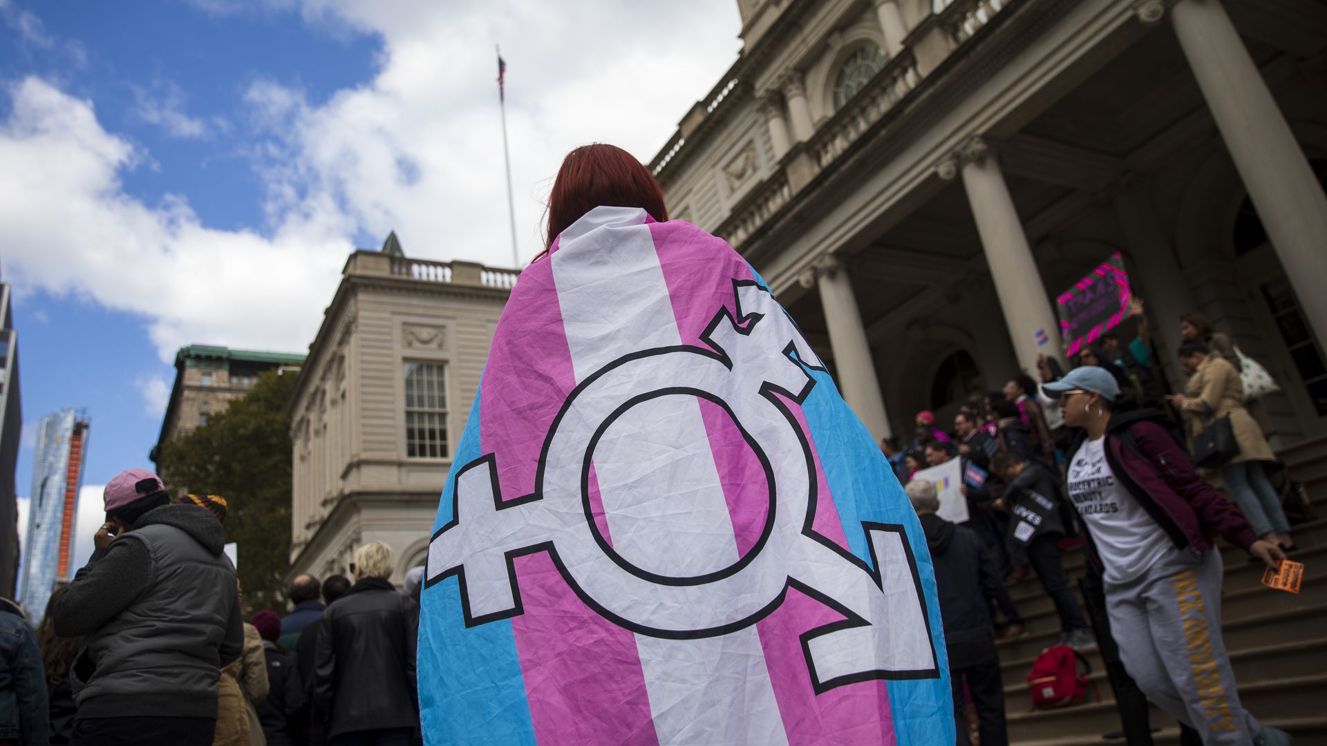 In this image, a person with red hair carries a transgender pride flag around their neck like a cape