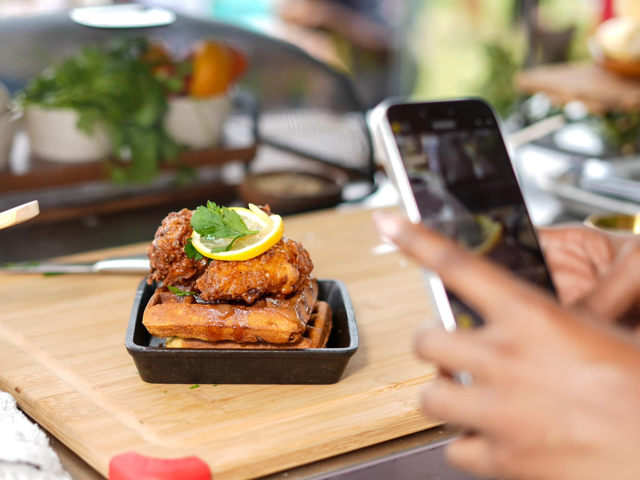 Close-up of a fried chicken and waffles with a lemon slice and parsley, served on a black tray. In the background a person holds a smartphone to photograph the dish.