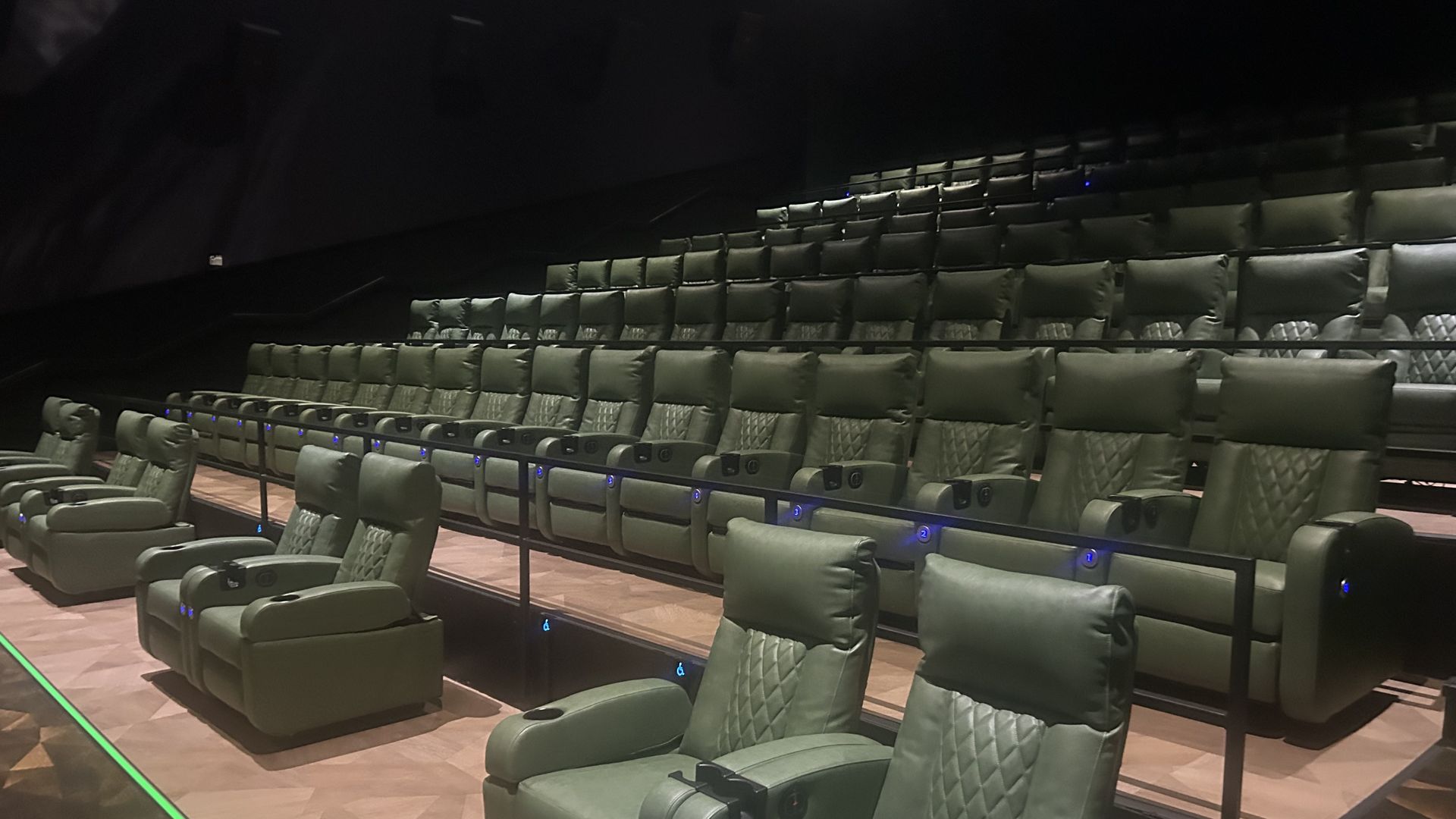 Dimly lit movie theater with many green cushioned reclining seats in curved, multi-tier rows; blue aisle lights along the floor.
