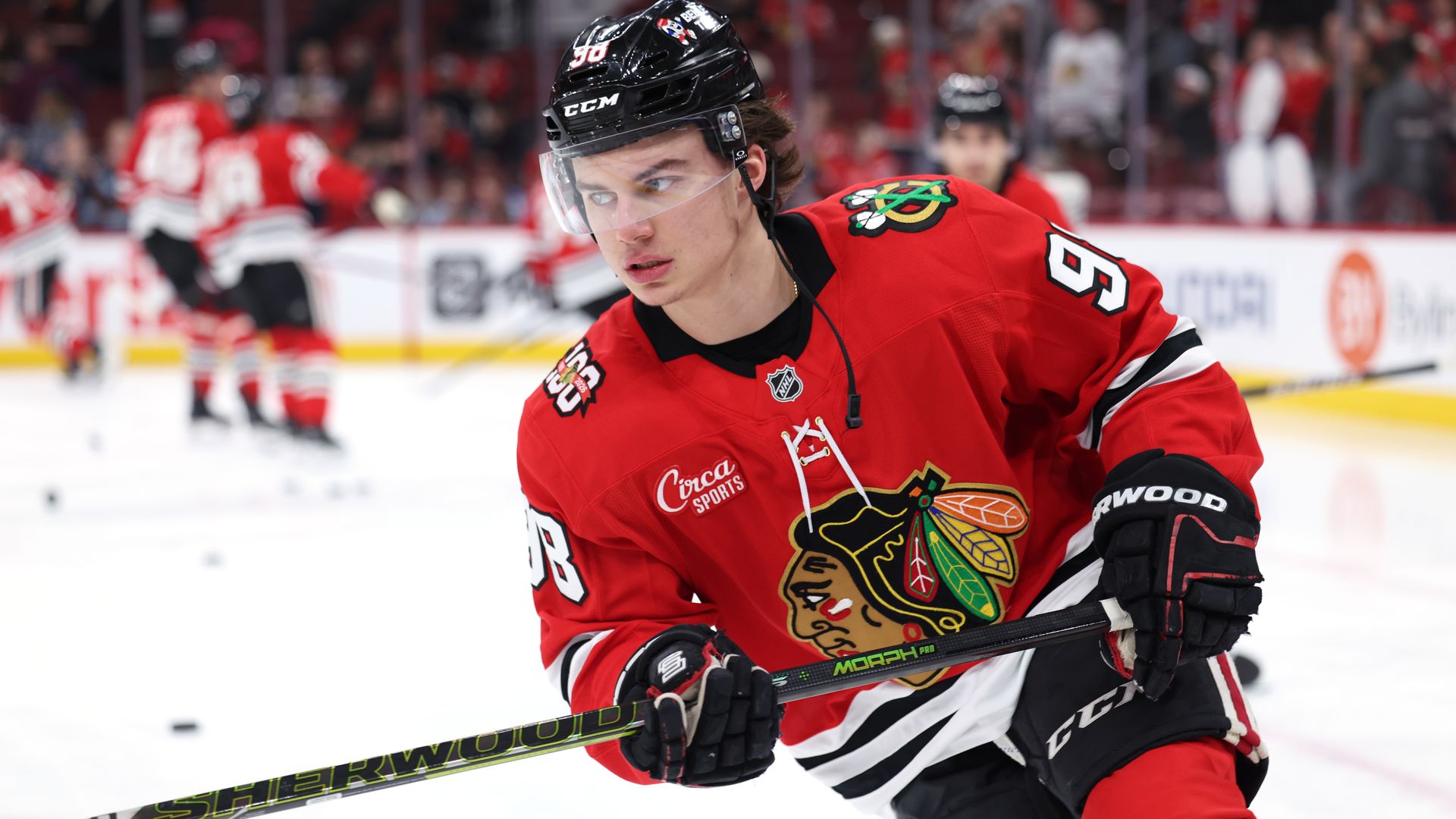 Hockey player in red Chicago Blackhawks jersey with number 98 and black helmet is skating on ice, holding a Sherwood hockey stick, with blurred players and crowd in the background.
