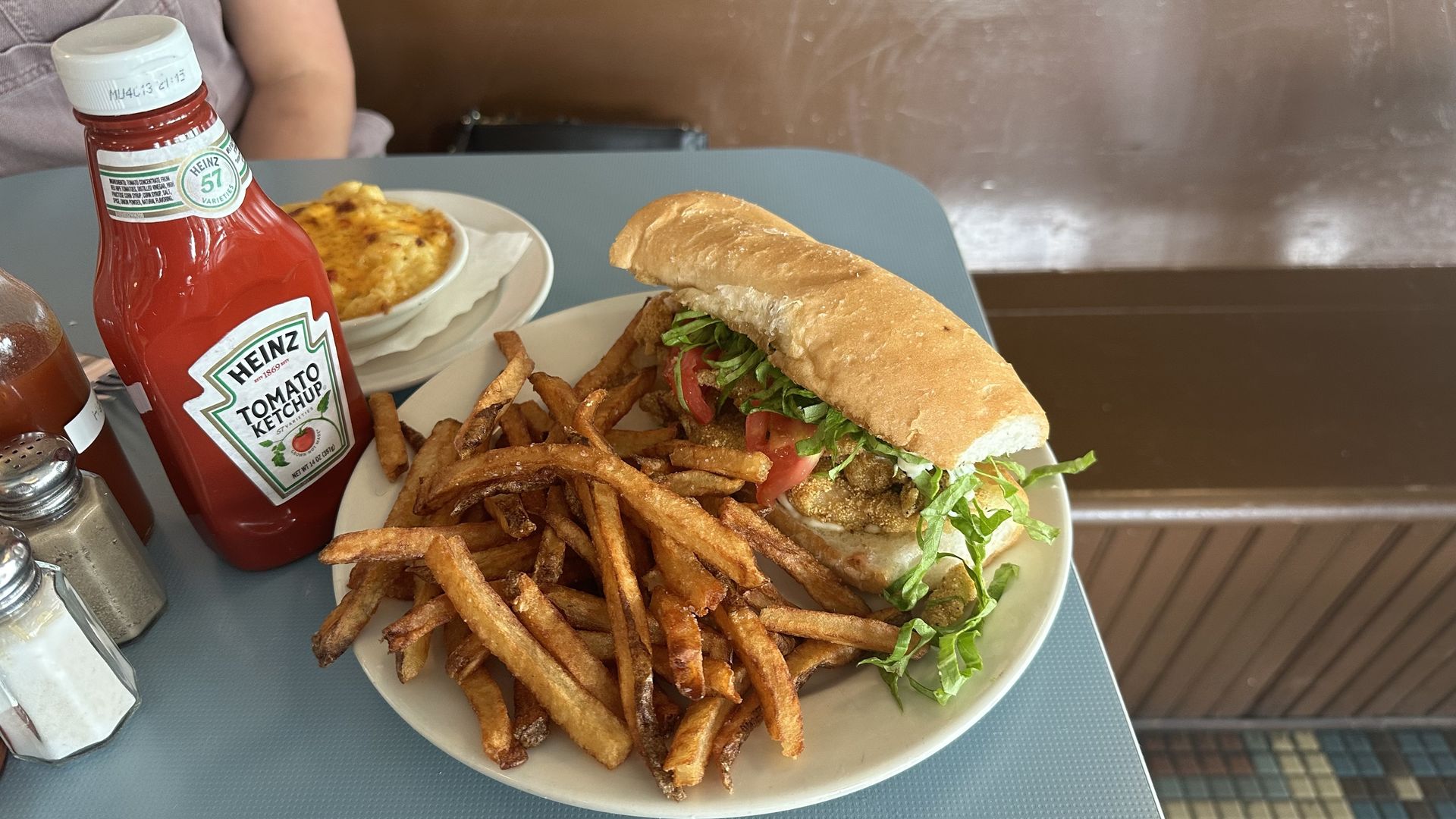 A blue diner-style table is set with salt, pepper, ketchup and a plate filled with fries and a catfish poboy. A small play of mac and cheese sits to the site.