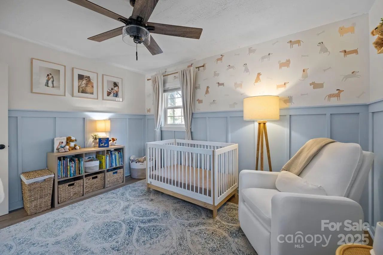 Bright nursery with light blue wainscoting, dog-themed wallpaper, white crib, white armchair with beige throw, bookshelves, and floor lamp under ceiling fan.