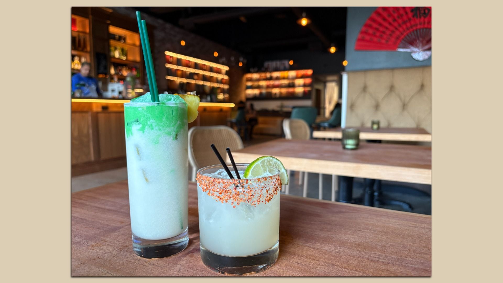 Two drinks on a wooden table in a dimly lit bar: a tall green and white layered frozen drink with a pineapple garnish and two green straws, and a short clear drink with a chili-rimmed glass and lime wedge.