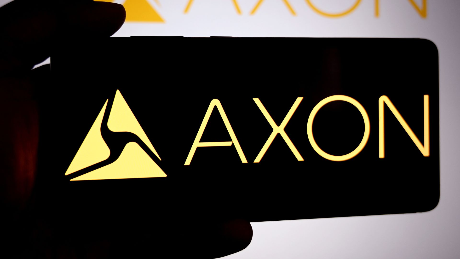 Silhouette of a hand holding a black smartphone in front of a bright display featuring the yellow AXON logo with a triangular emblem and the word AXON.