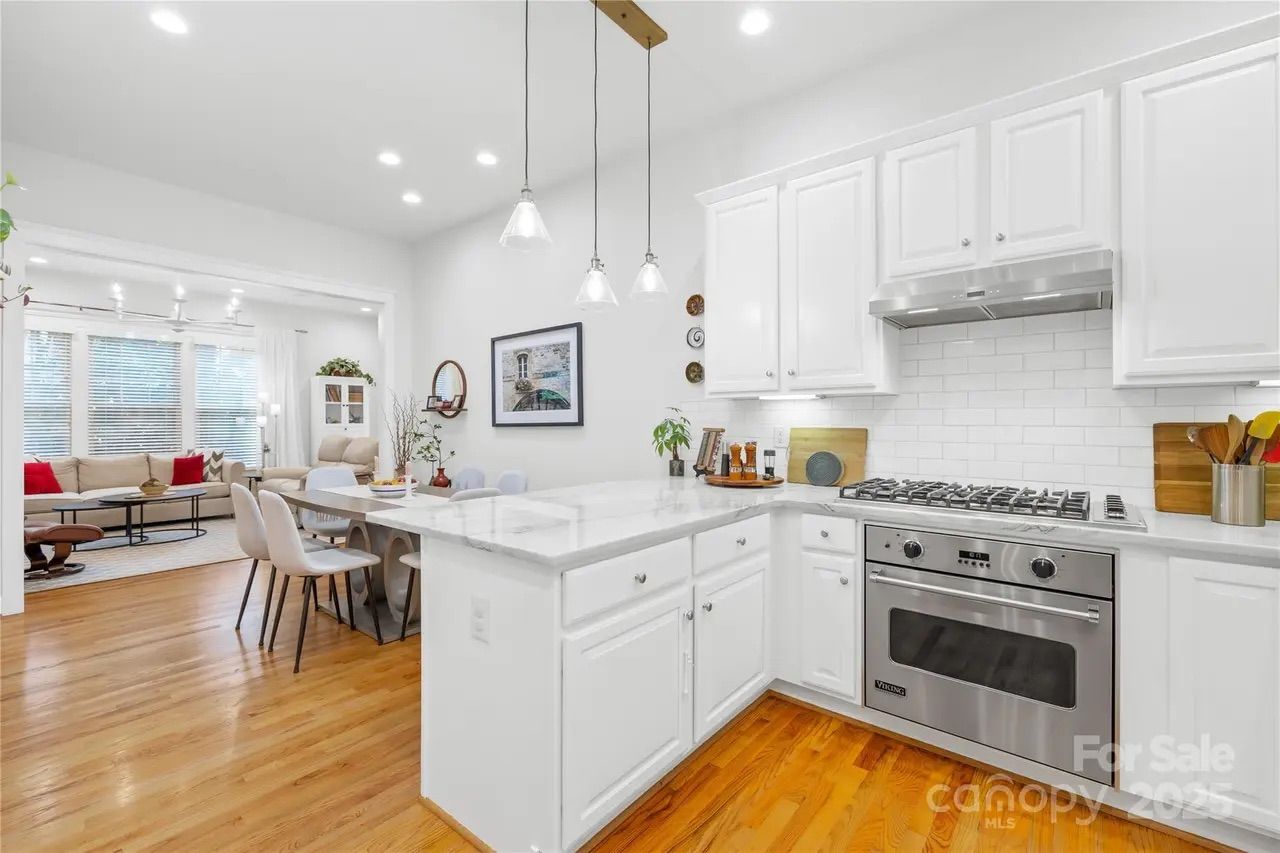 Bright white kitchen with marble countertops, stainless steel Viking stove, white cabinets, wooden floors, and hanging pendant lights. Adjacent living area with beige furniture and red pillows.